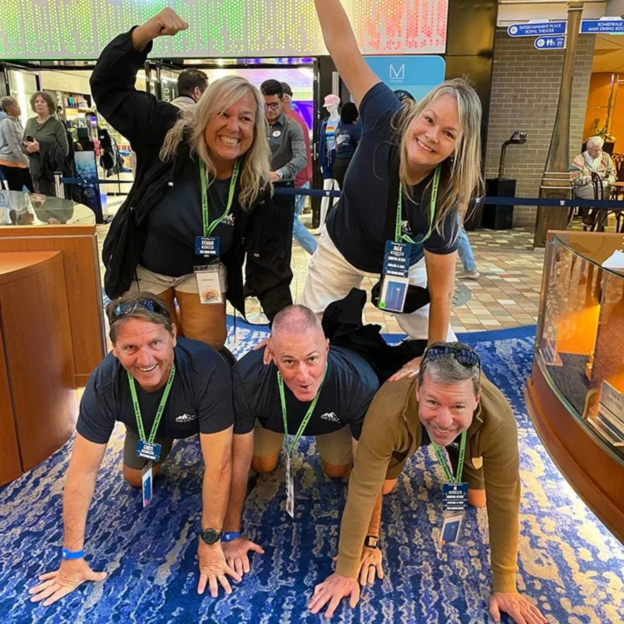 Two women standing on their hands on a blue patterned carpet in what appears to be an airport, smiling and raising their arms, with other people and signs in the background.
