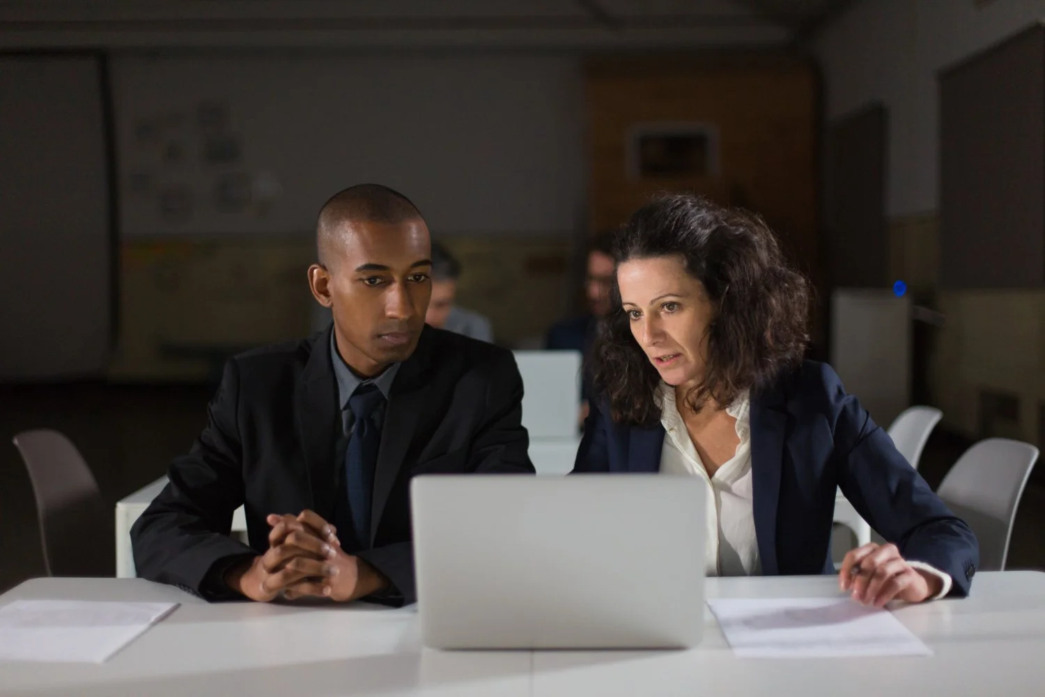 Two professionals, a man and a woman, looking at a laptop on a table in a meeting room with papers and a dark background.
