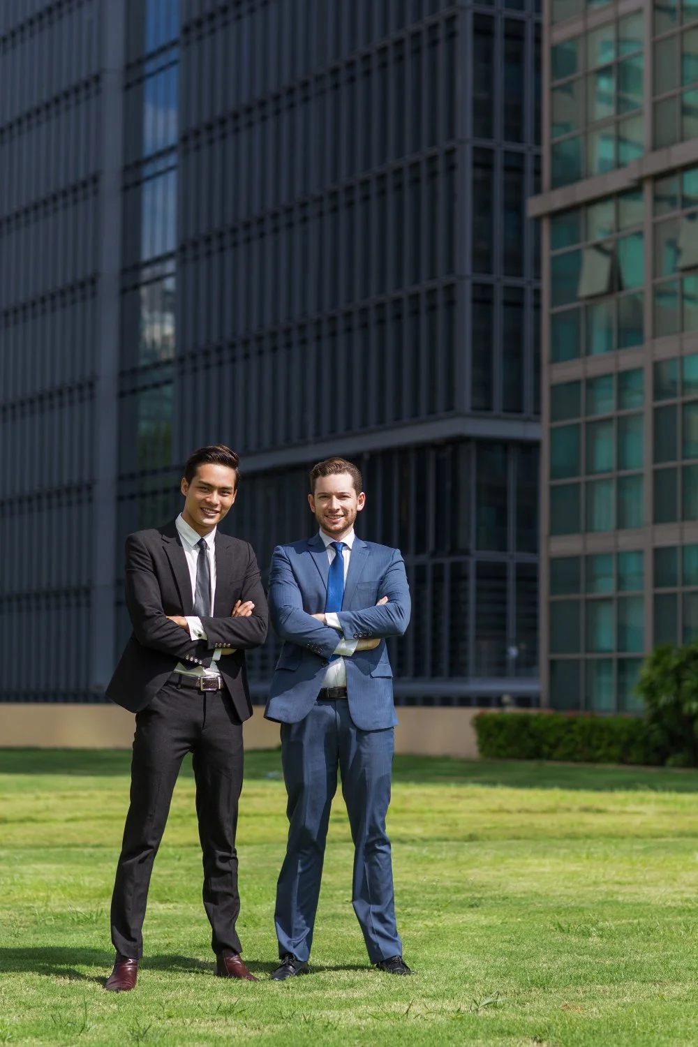 Two smiling businessmen in suits with arms crossed, standing outdoors on a grassy area in front of a modern glass office building.