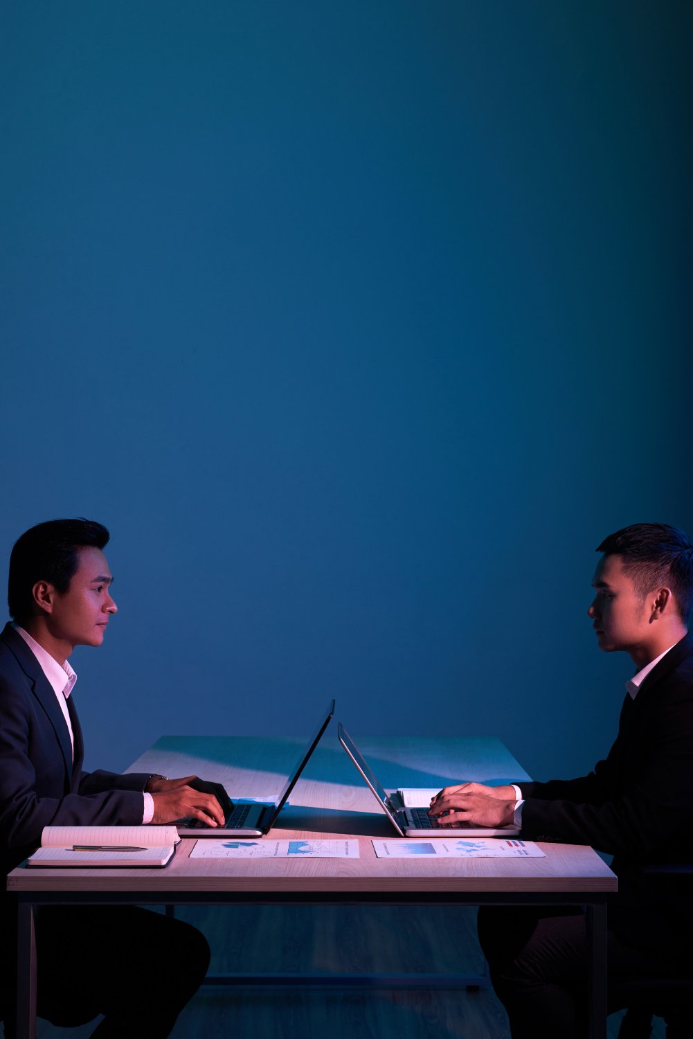 Two men in business suits sitting at a table, facing each other, working on laptops in a dimly lit room with blue background.