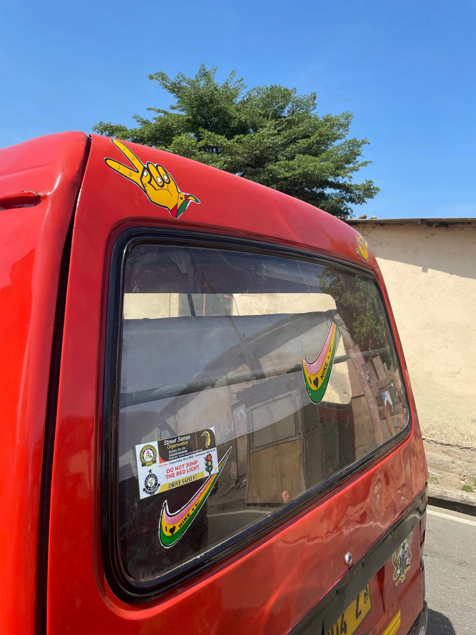 Red vehicle with Nike and hand peace sign stickers on the window and body, parked outdoors under a blue sky with a tree and wall in the background.