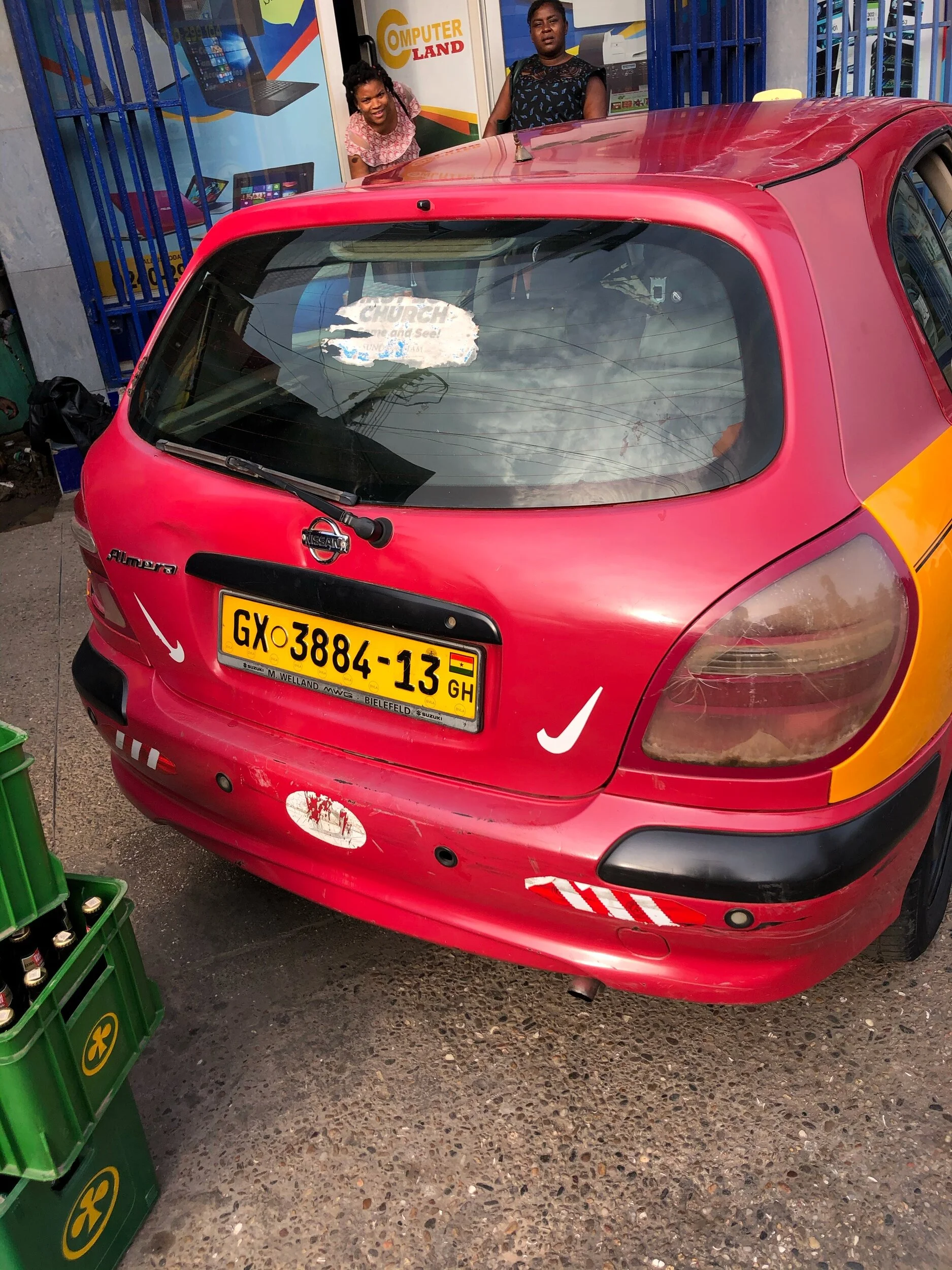 The image shows the rear view of a red Nissan Almera car parked on a street. The car has a yellow and black Ghanaian license plate with the registration GX 3884-13. The back of the car has some stickers, including a white Nike swoosh. Two women are s