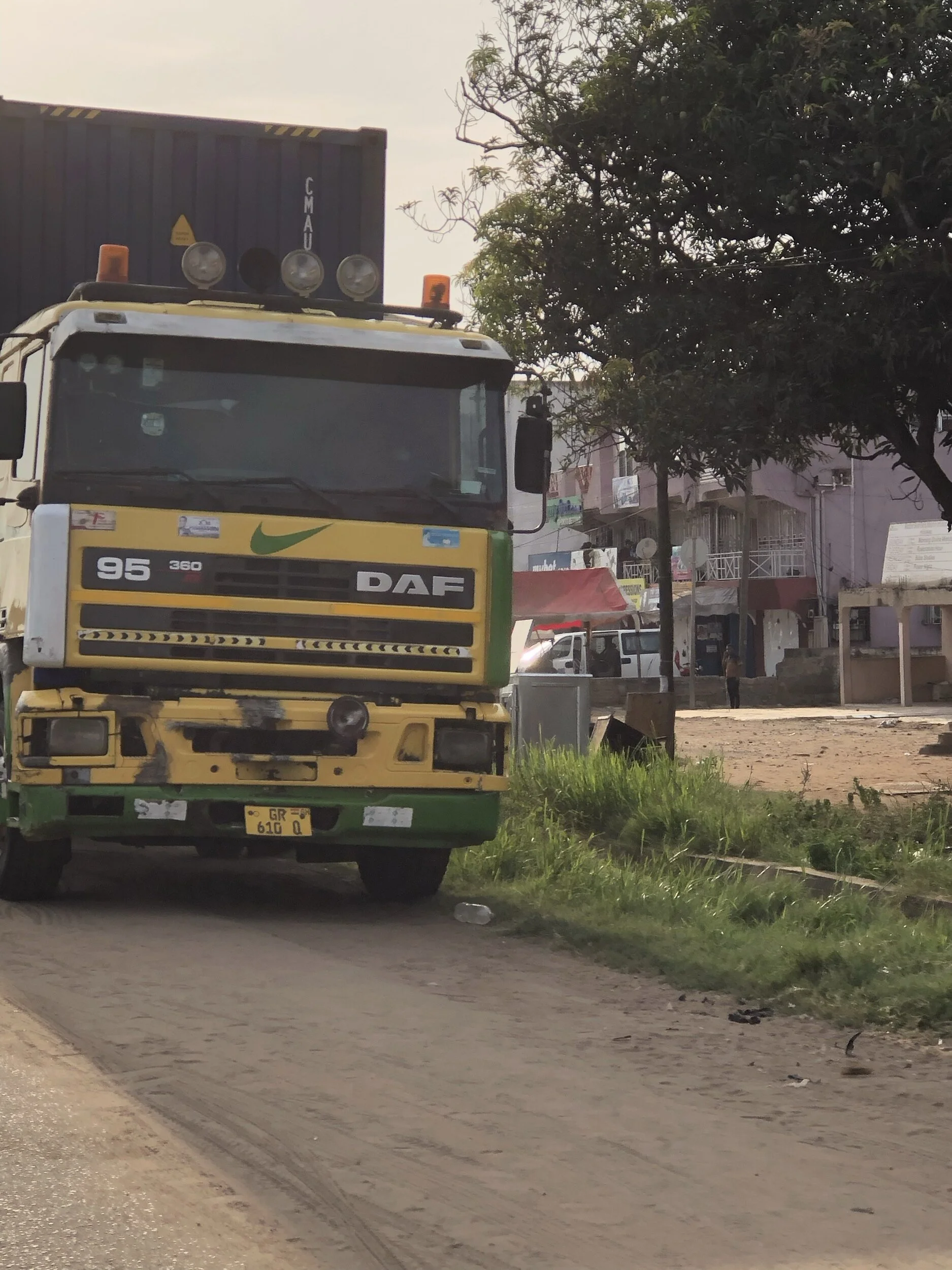 A yellow and green DAF truck parked on a dirt street with buildings and trees in the background.
