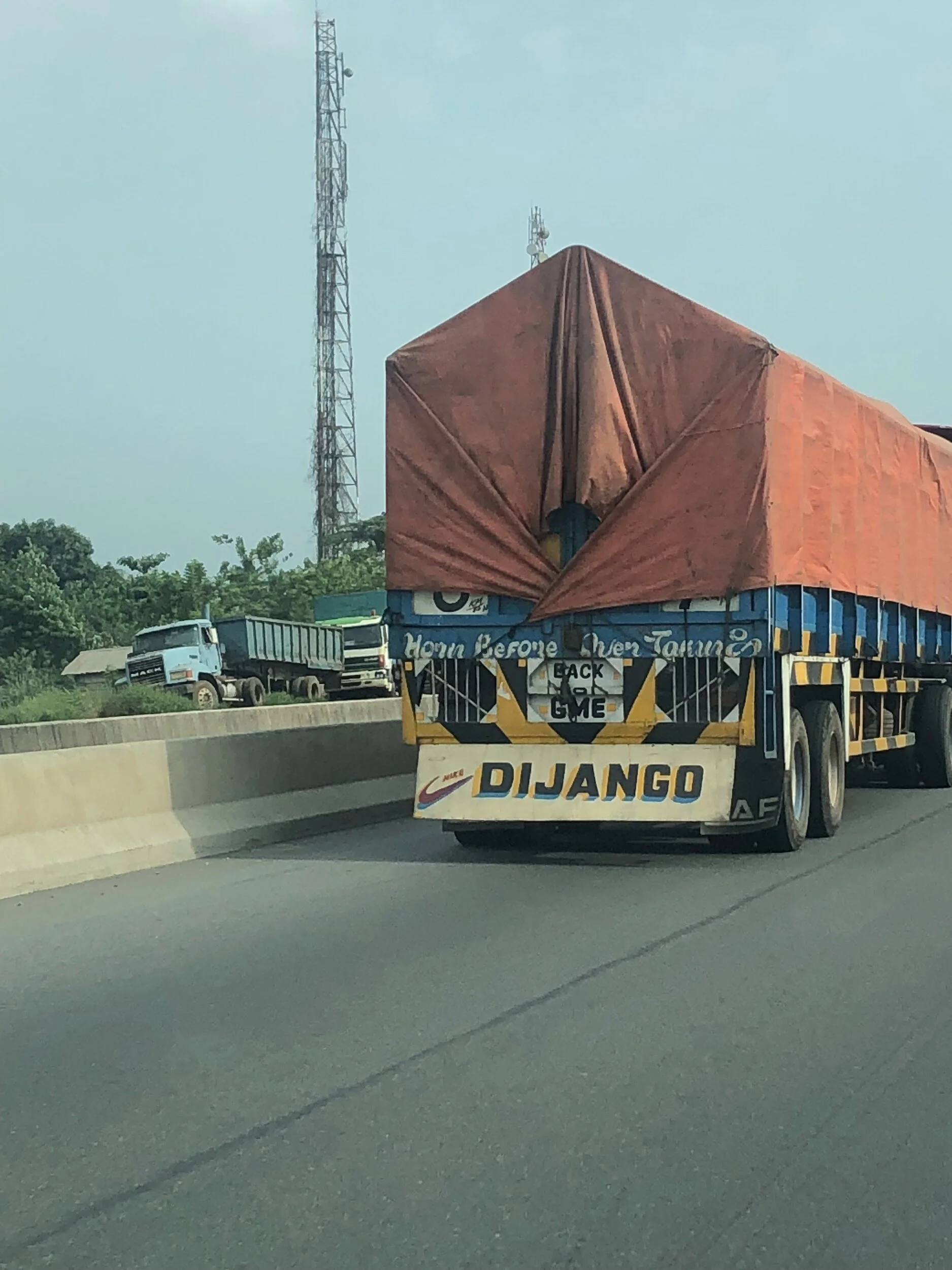 A large truck on a highway with a red tarp covering its cargo. The back of the truck has the words 'Django' and a sign that reads 'Back 2 Game.'