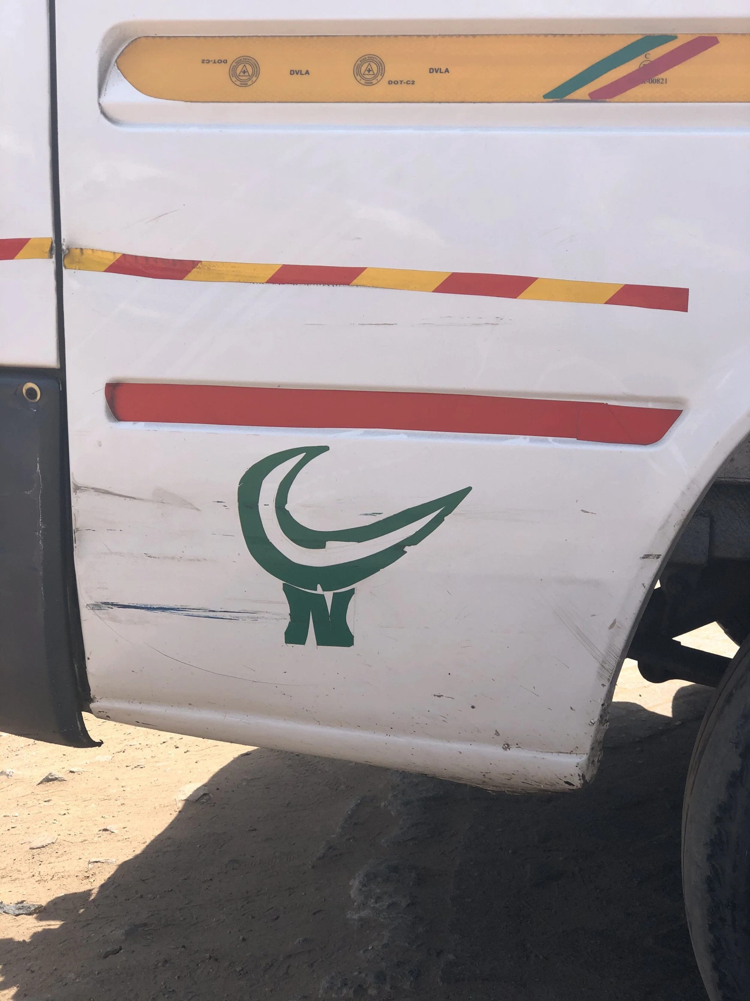 Close-up photo of a white vehicle's side, featuring a green Nematodes logo on the lower part, yellow and red reflective striping, and damage to the body with scratches and dirt.