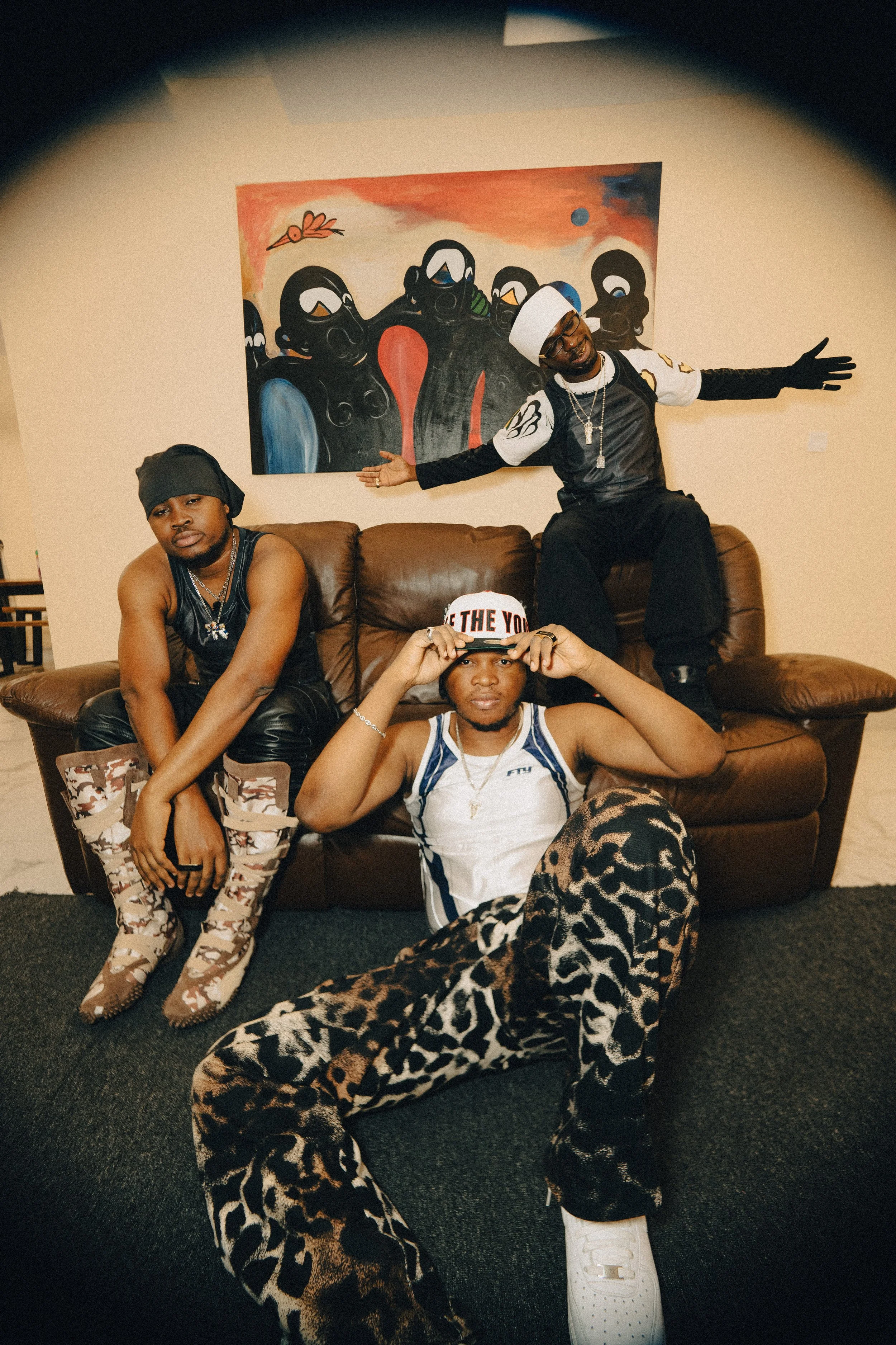 Three young men posing in a living room with a brown leather sofa and colorful artwork behind them. One man is seated on the floor wearing leopard print pants, white sneakers, and a white tank top, holding a cap. Another is sitting on the sofa wearin