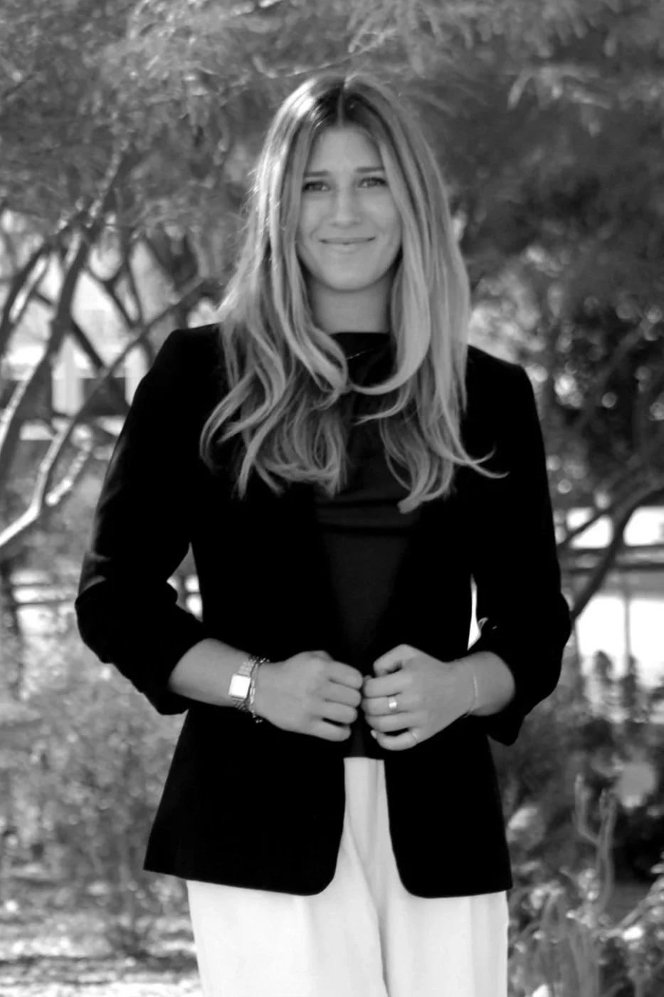 A woman with long, wavy brown hair and light makeup, wearing a black top, poses against a marble wall background.