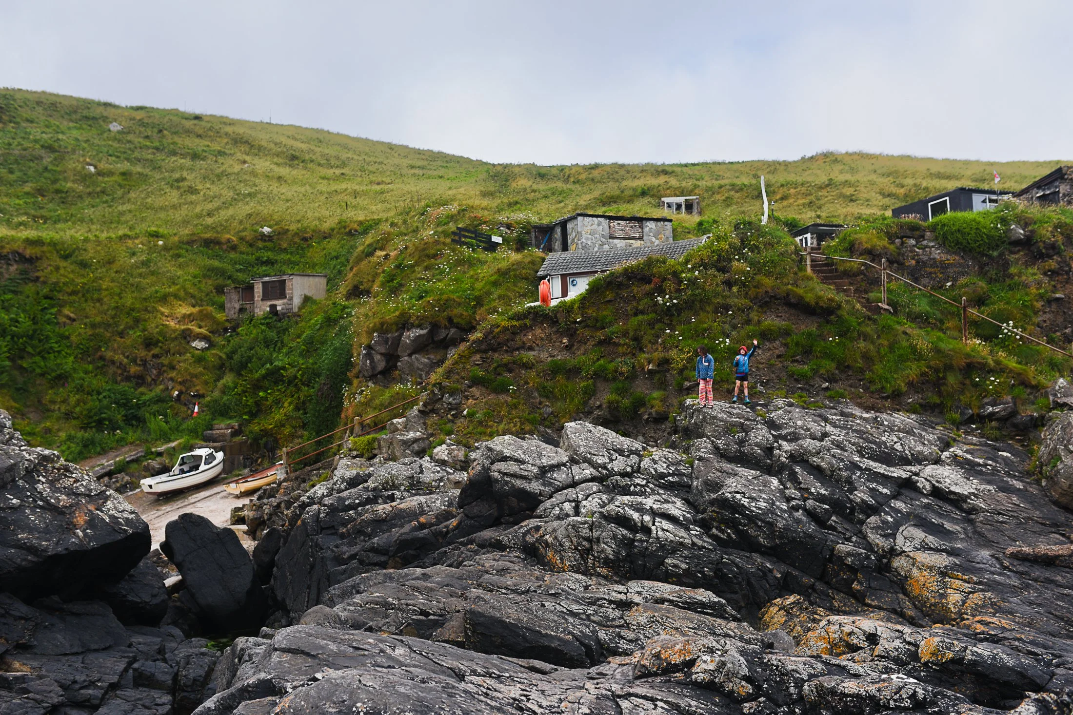 A Cornish cove with rocks in the foreground and two tiny figures stand on them. A fishing boat to the left and then green cliffs and huts behind them to a blue cloudy sky. The photo gives context by showing the size of the figures in the landscape.