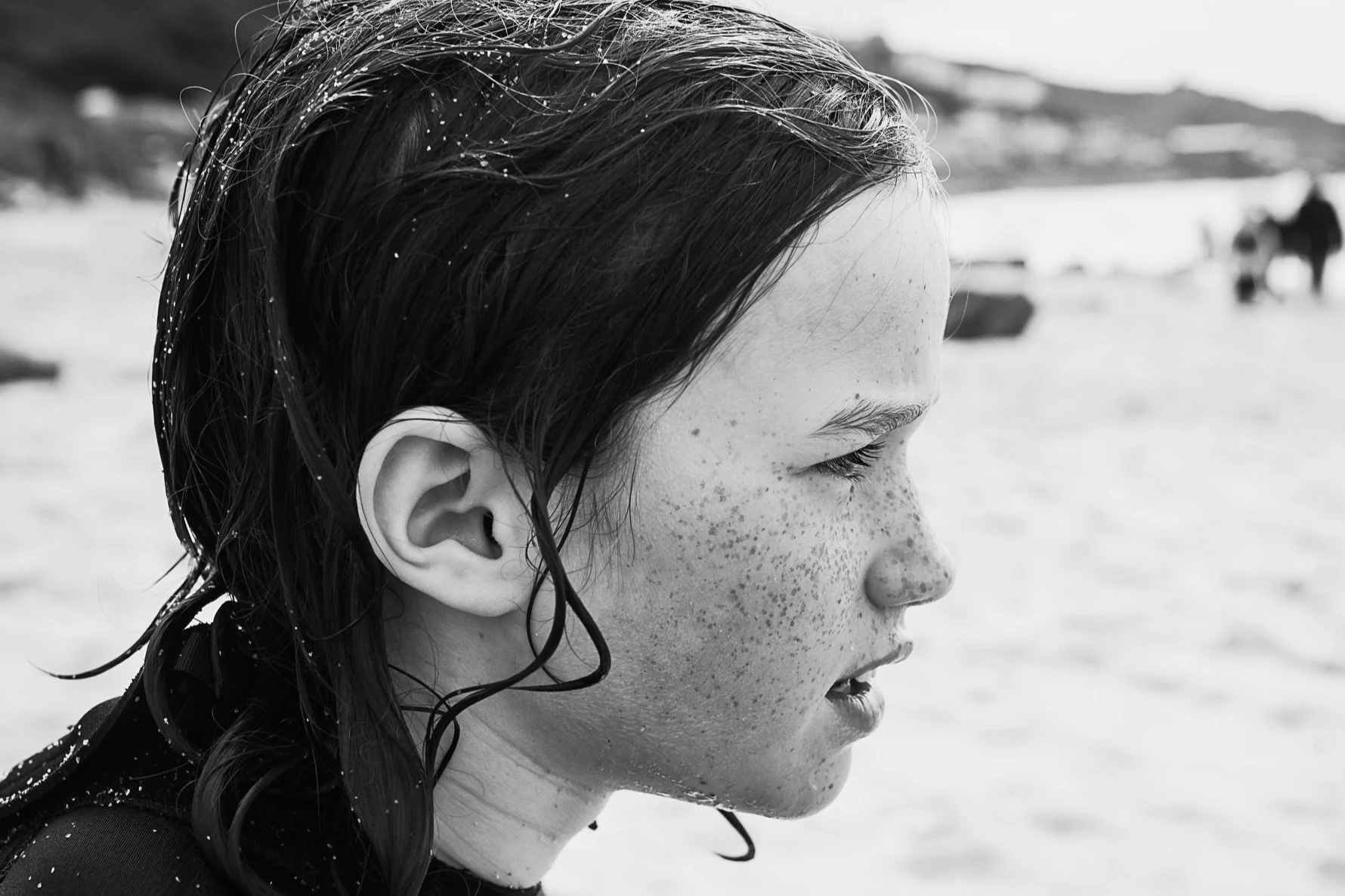 A close up black and white portrait of a ten year old in profile. There are freckles in on their cheeks and their hair is wet and sandy. Their face looks thoughtful and unaware of the photographer.
