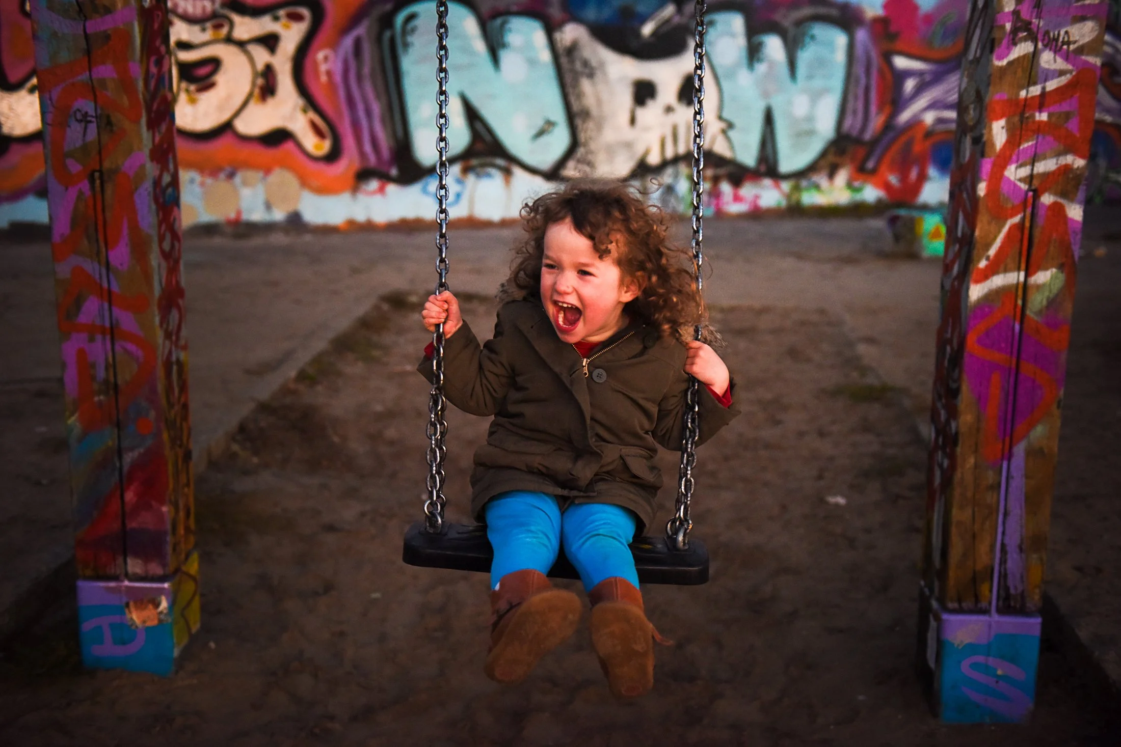 A three year old child with curly shoulder length hair is on a swing at dusk in an urban environment with graffiti all around. The word NOW is behind them with the O as a skull. The child has her mouth open screaming.