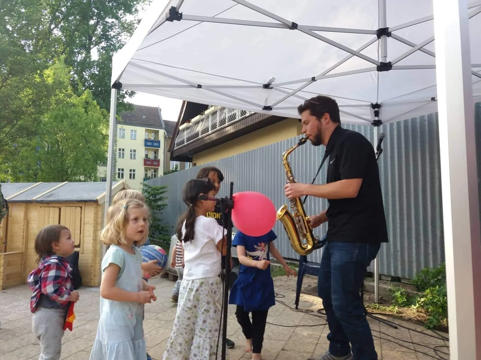 Ein Mann, der Saxophon spielt und draußen unter einem weißen Sonnendach für Kinder auftritt, während diese ihm zuschauen und vor ihm spielen.