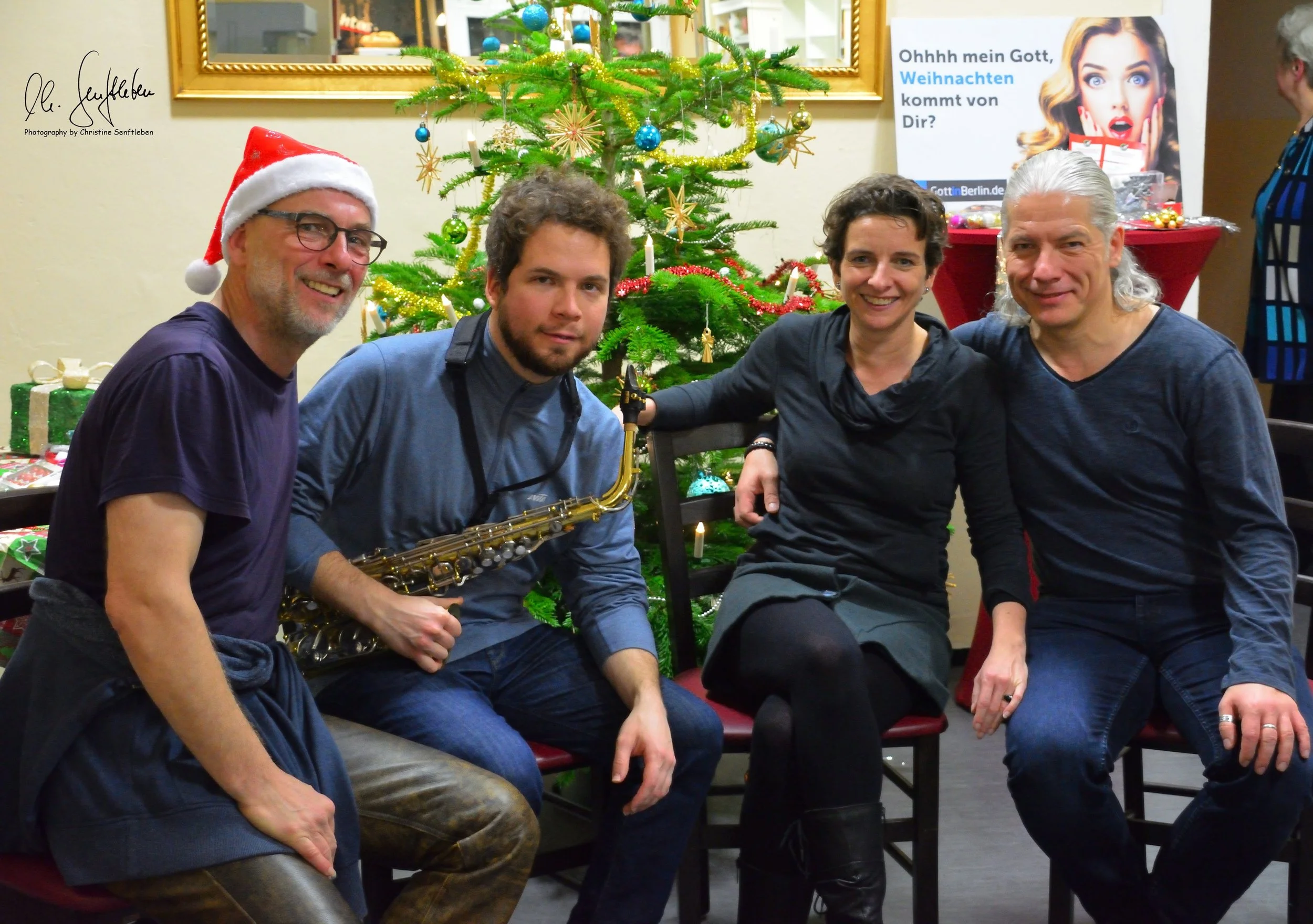 Four people smiling and sitting in front of a decorated Christmas tree; Klim is holding a saxophone and wearing a Santa hat.