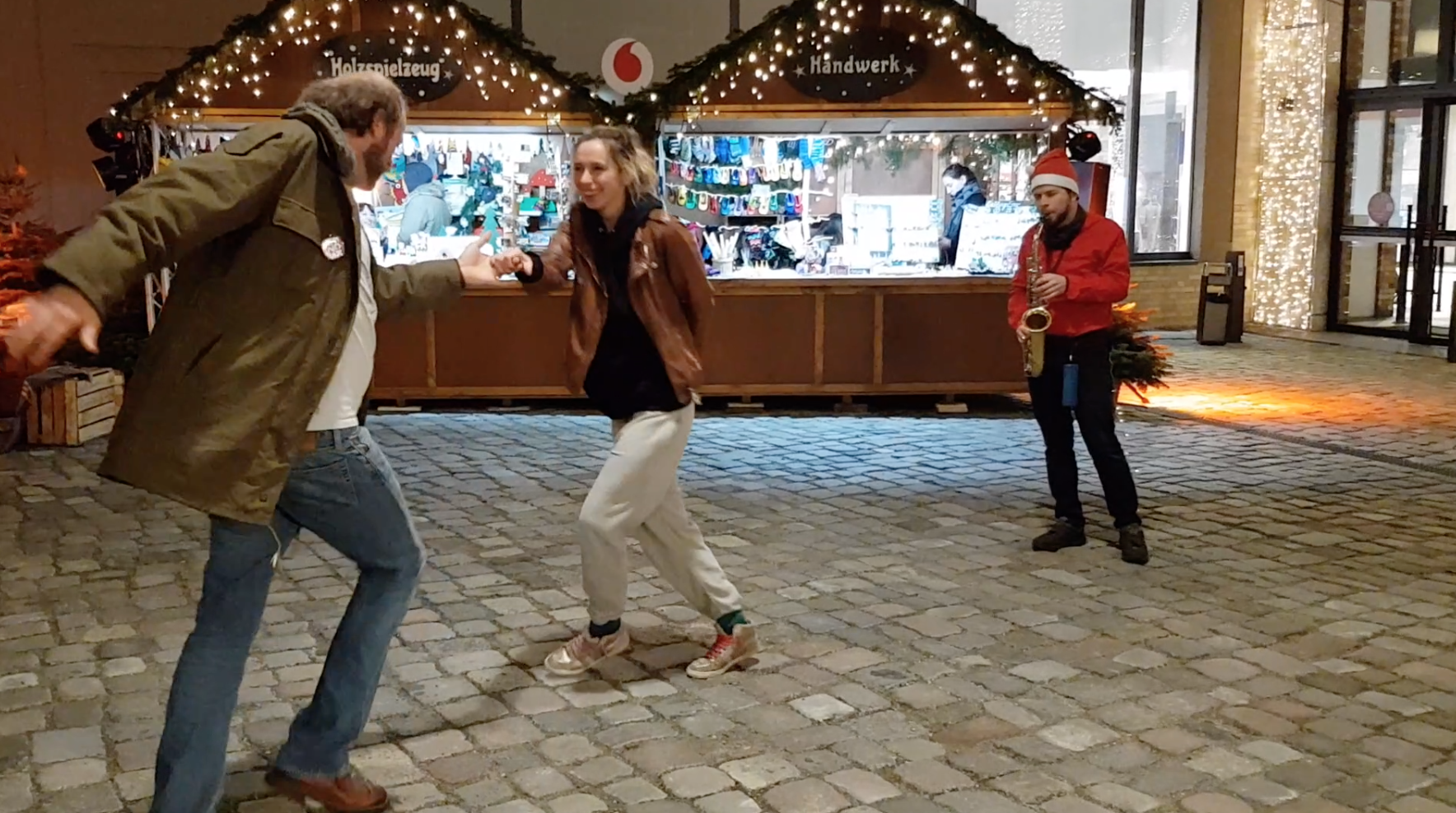 Two people dancing in front of Christmas market stalls with lights, accompanied by a saxophone player wearing a Santa hat.