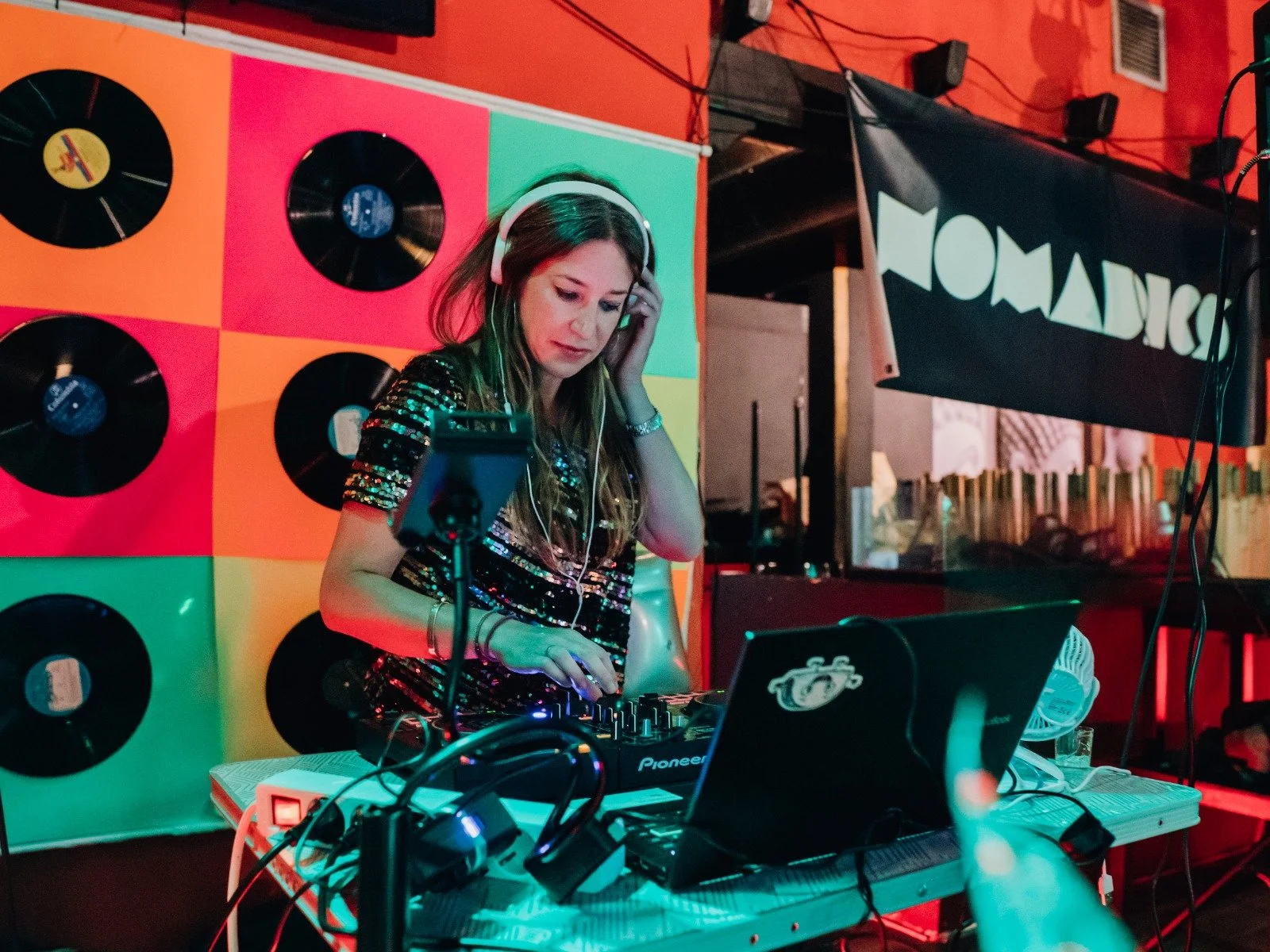 DJ playing music with turntables and laptop in colorful setting, with vinyl records on the wall.