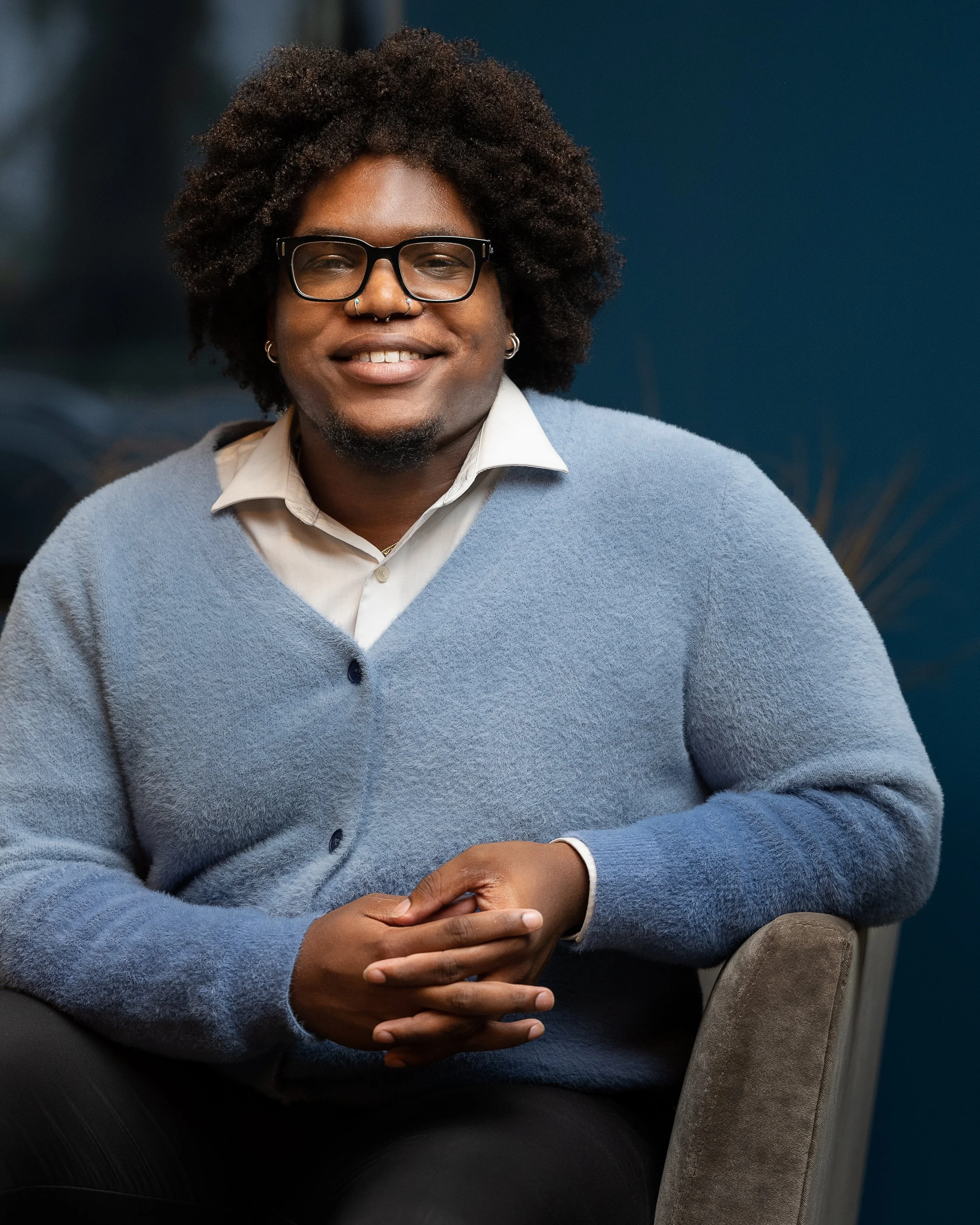 Corporate headshot of a smiling black man with curly hair, glasses, and a septum piercing, sitting in a chair against a blue background, wearing a light-colored shirt and a blue cardigan.