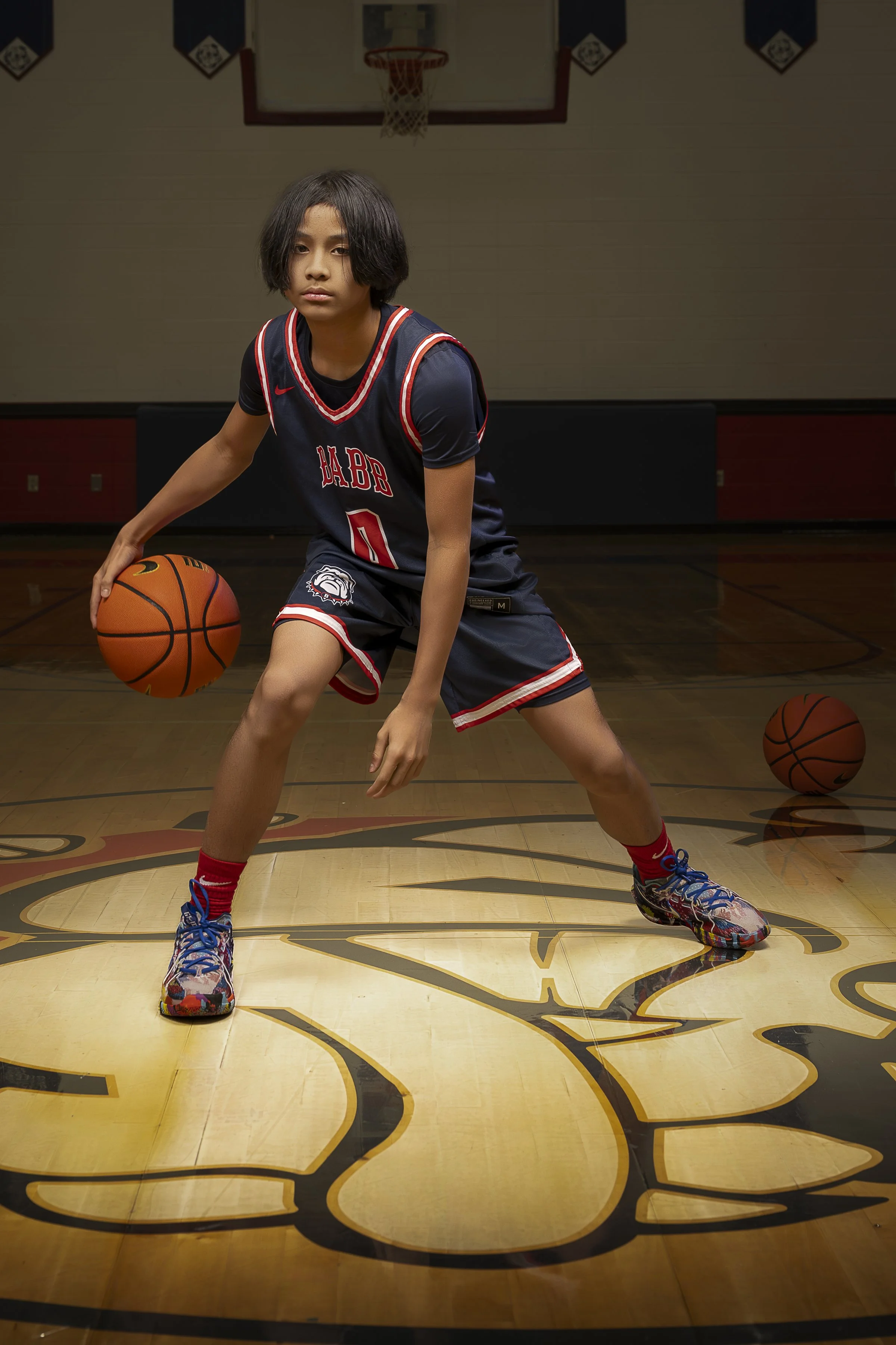 A young basketball player in a navy blue and red uniform practicing dribbling on an indoor court.