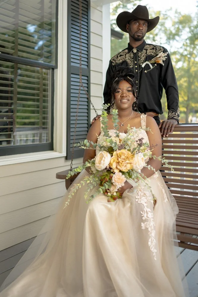 Wedding photography portrait. A woman in an elegant dress holding a bouquet, sitting on a porch swing, with a man standing behind her wearing a cowboy hat and embroidered shirt.