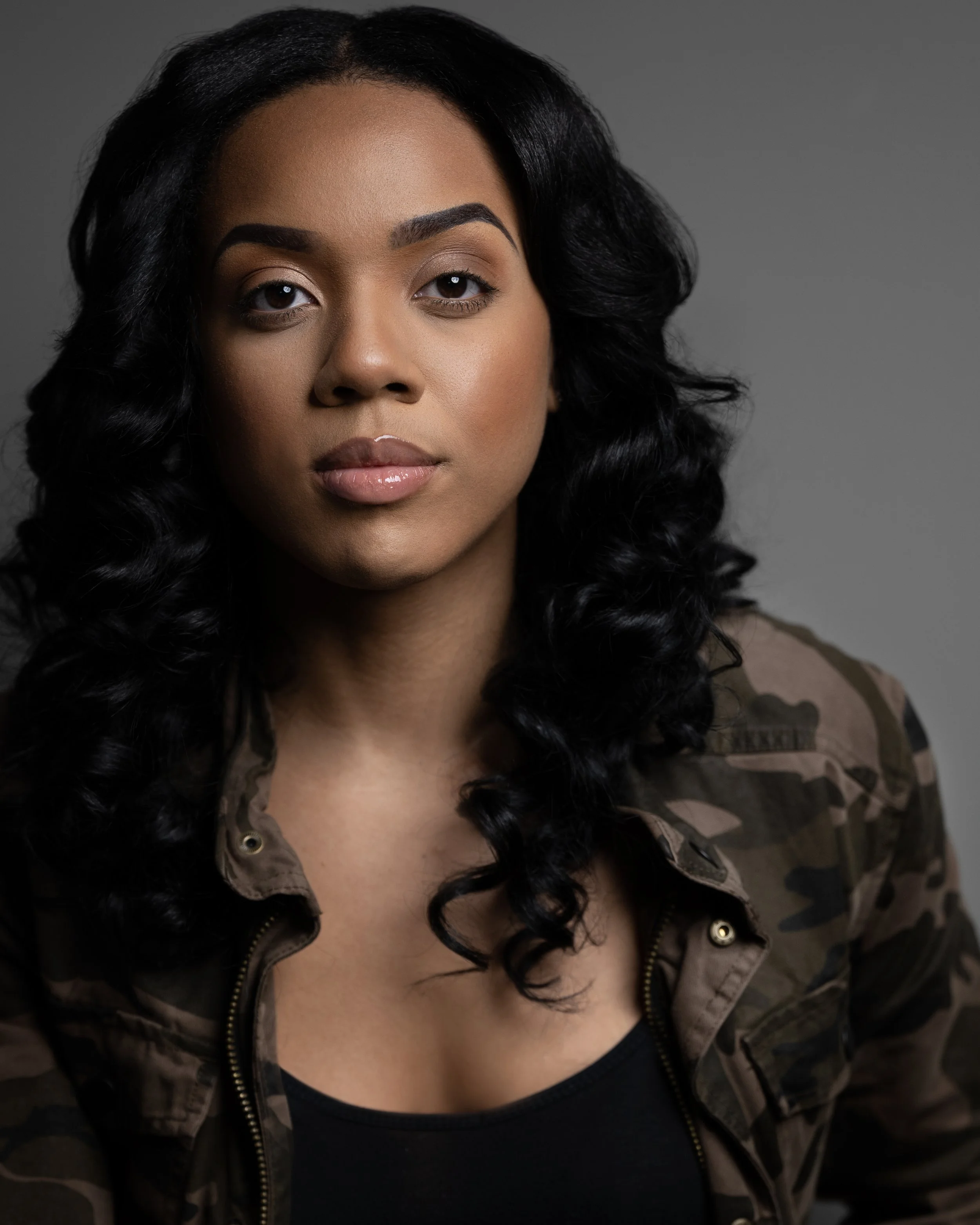 A woman with long curly black hair, tan skin, and wearing a camouflage jacket over a black top, looking directly at the camera against a gray background.