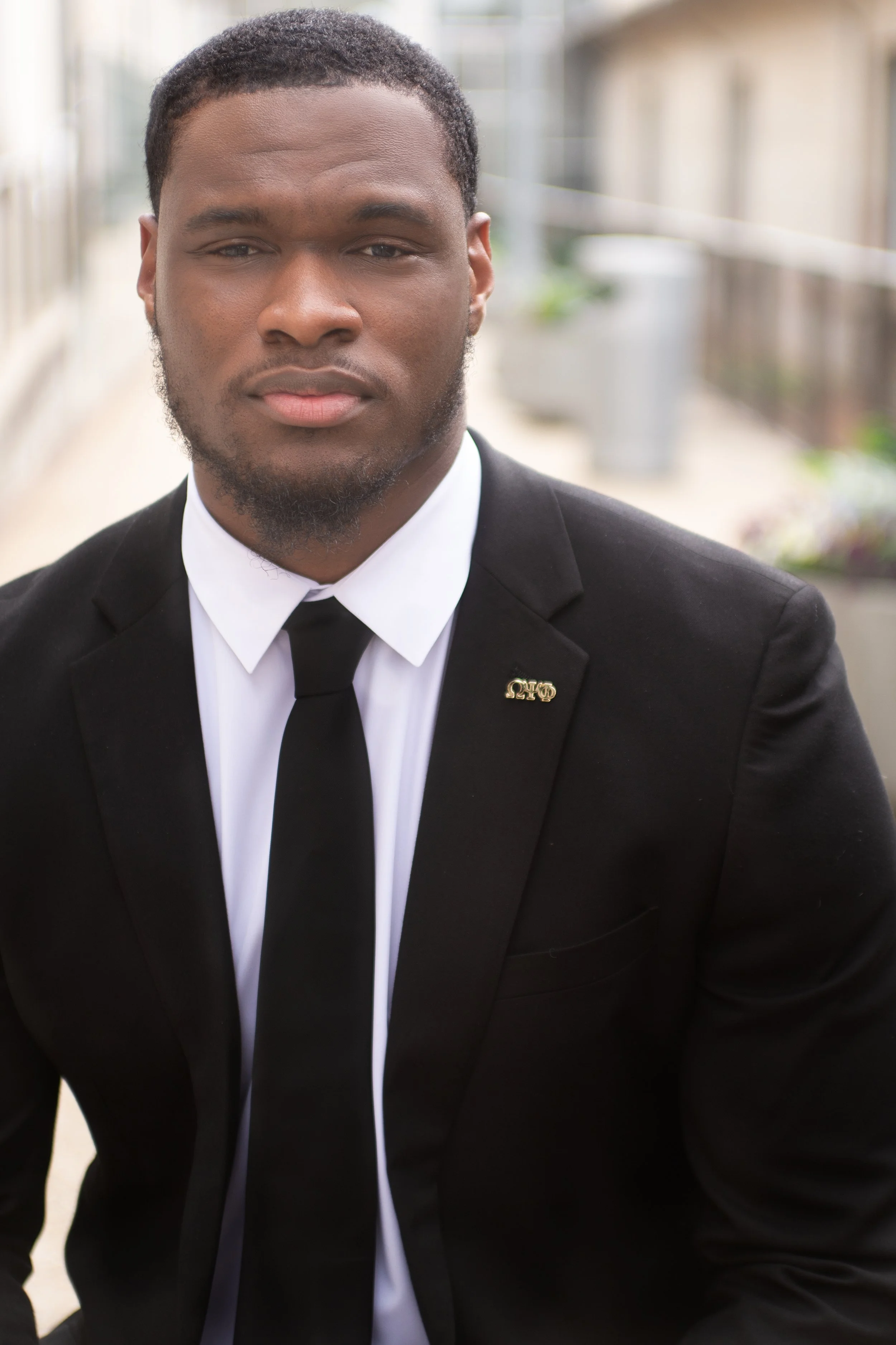 Corporate headshot of a young professional man dressed in a black suit, white shirt, and black tie, standing outdoors with a blurred background.