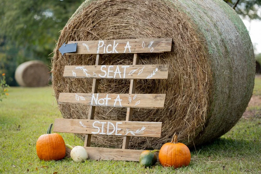 Wooden sign in front of hay bale with pumpkins, stating "Pick a Seat, Not a Side."