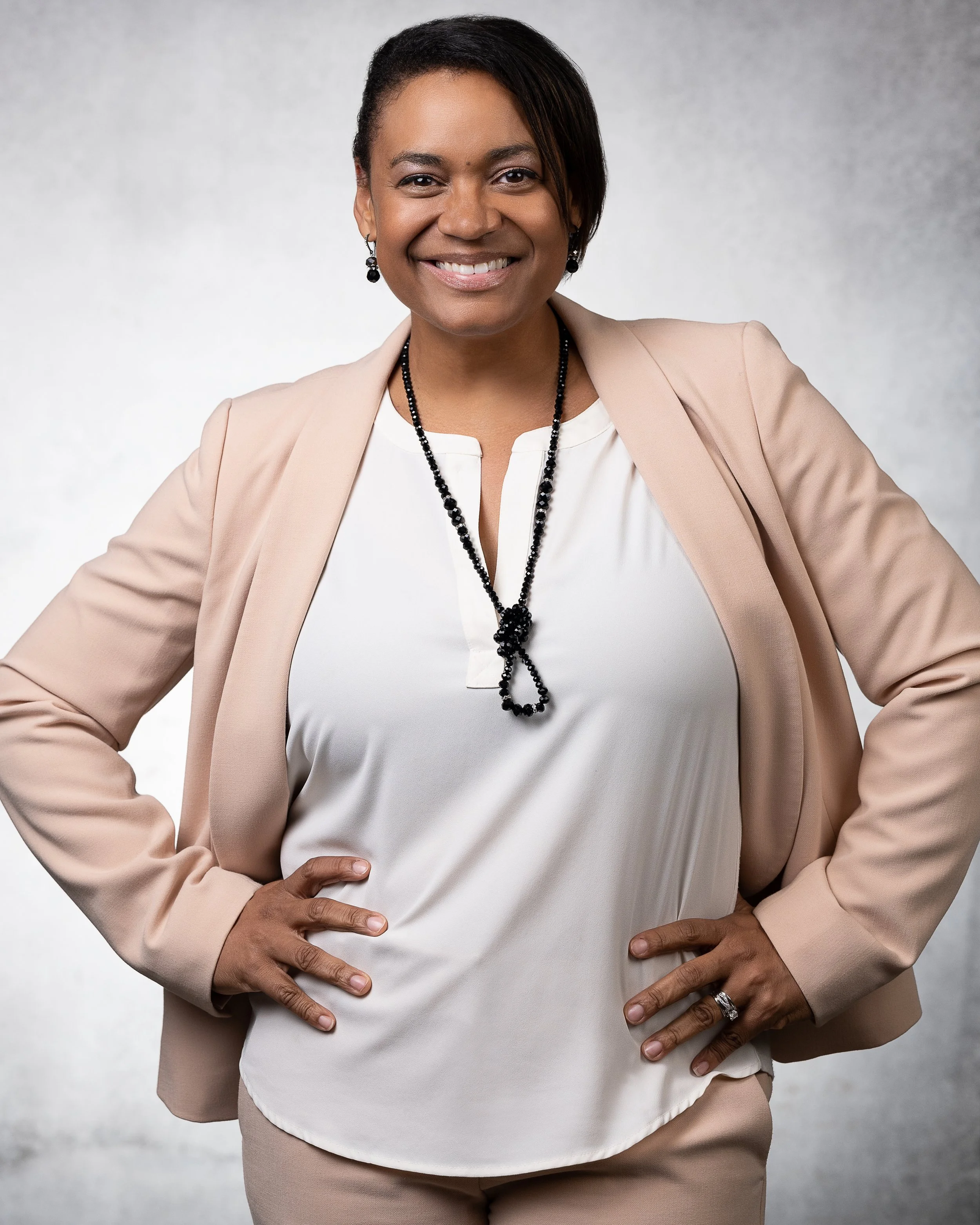 Corporate headshot of a smiling woman with short dark hair wearing a beige blazer, white blouse, and black necklace, standing with hands on hips against a plain background.