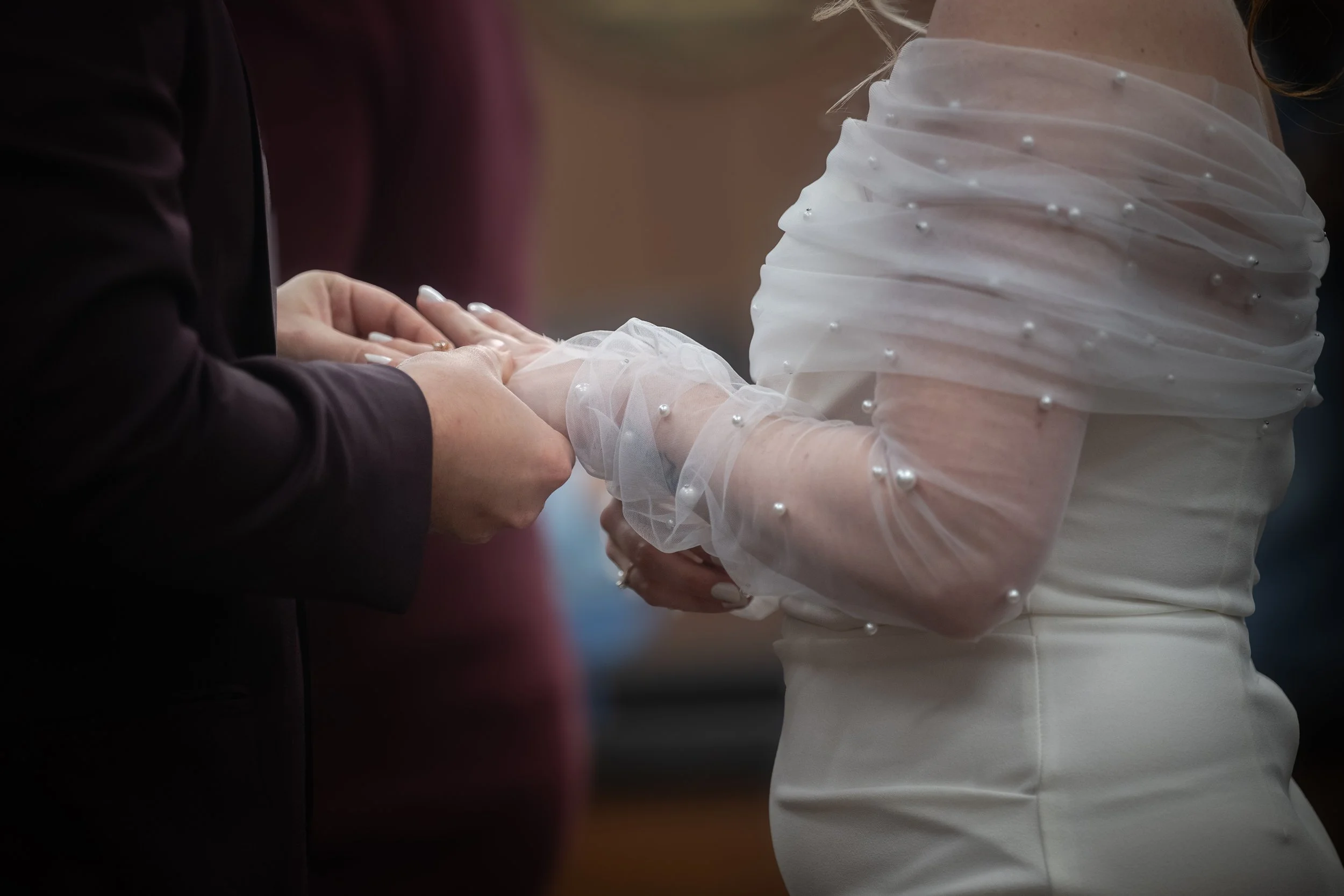 Close-up of a couple exchanging rings during a wedding ceremony. One person is wearing a white dress with pearl details, and the other is in a dark suit.