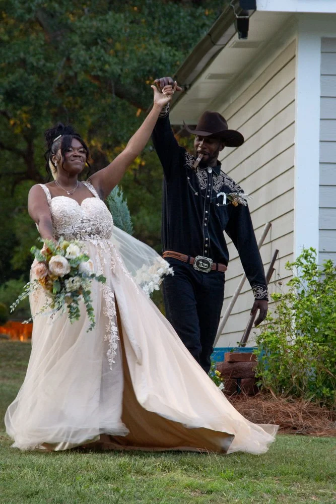 A bride in a white wedding dress holding a bouquet, and a groom in a cowboy outfit walking outdoors.