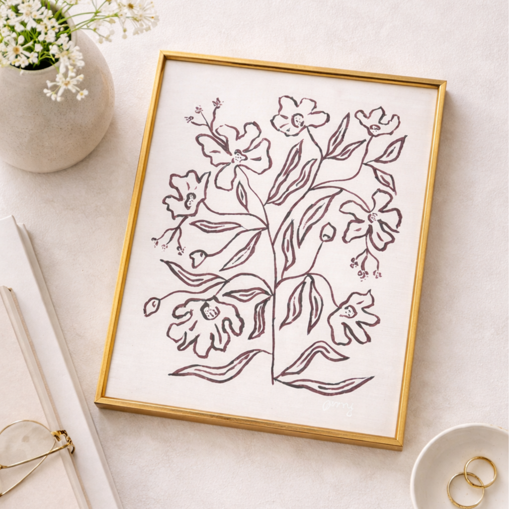 A framed line drawing of a floral design on white paper, placed on a light-colored surface. To the top left, there is a white vase with small white flowers. To the bottom left, are some white and gold items, possibly stationery or accessories. To the bottom right, there's a small white dish with two gold rings inside.