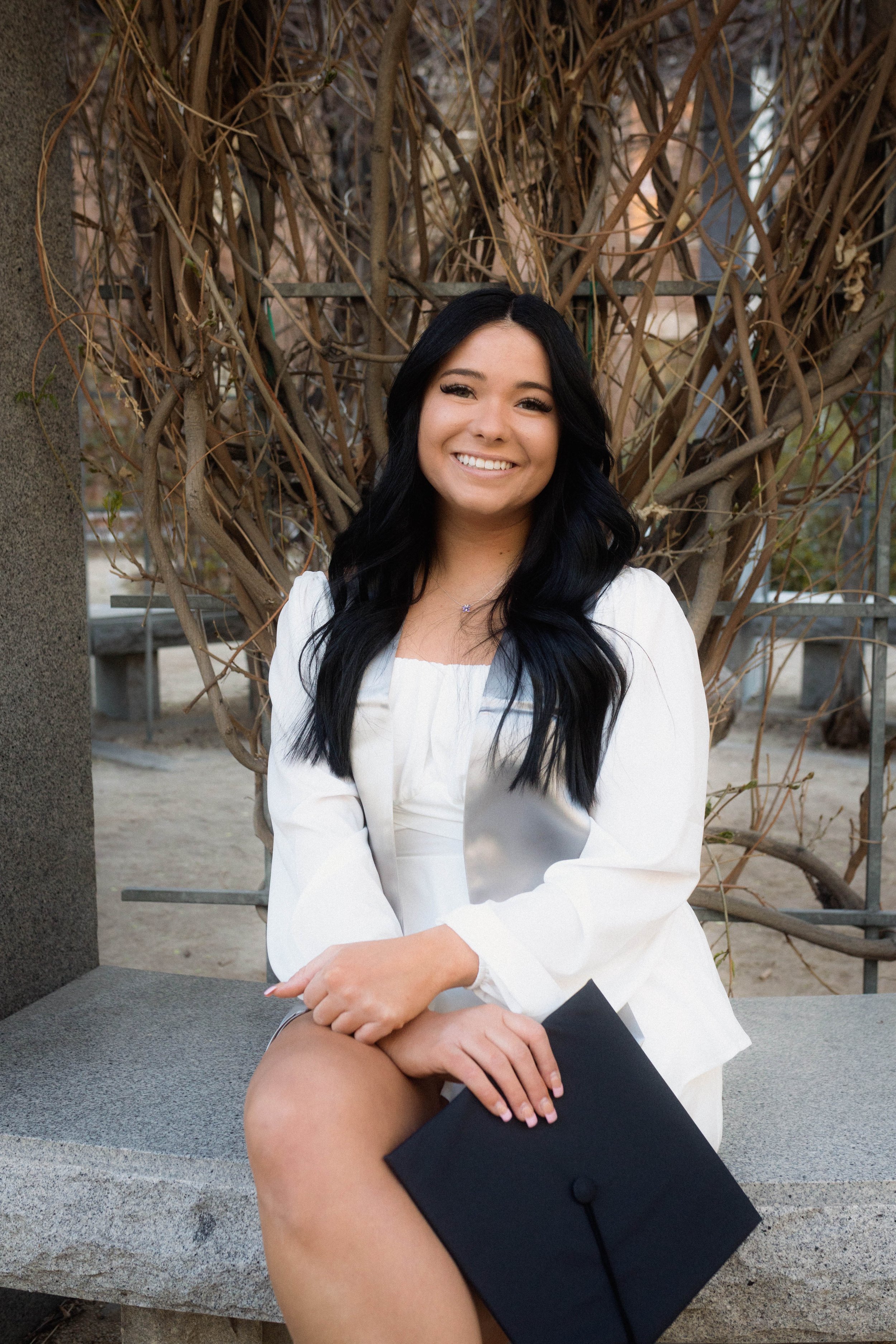 Young woman with long black hair, sitting on a stone bench, holding a graduation cap, smiling outdoors with leafless bushes behind her.