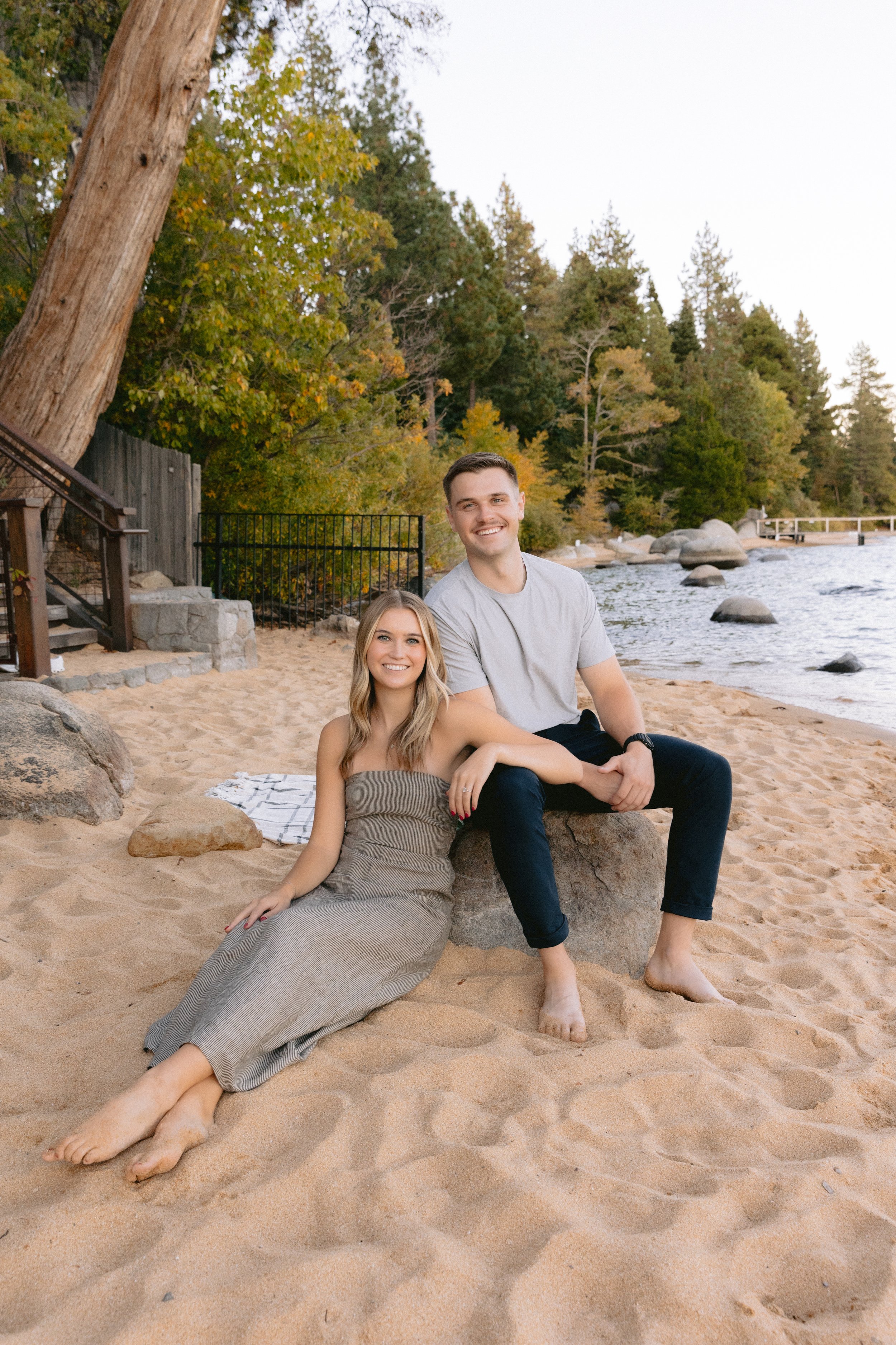 A smiling young couple sitting on a rock on a sandy beach with trees and water in the background