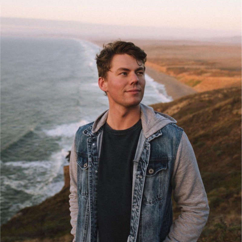 A young man with short brown hair in a denim jacket and black t-shirt standing on a cliff overlooking the ocean during sunset.