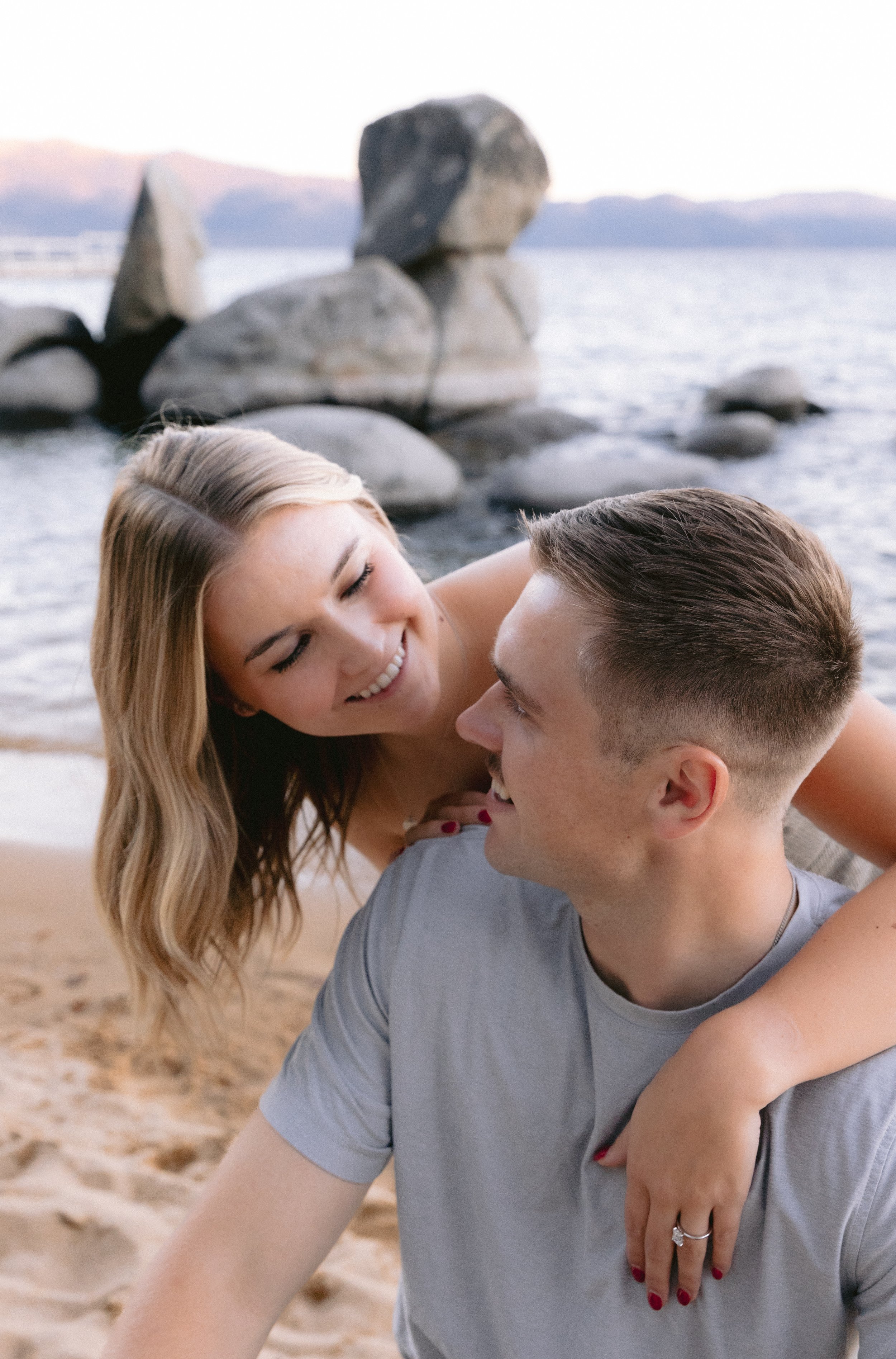 A smiling woman with long blonde hair is leaning on and looking at a man with short brown hair who is sitting on the sand by a lake with large rocks in the background.
