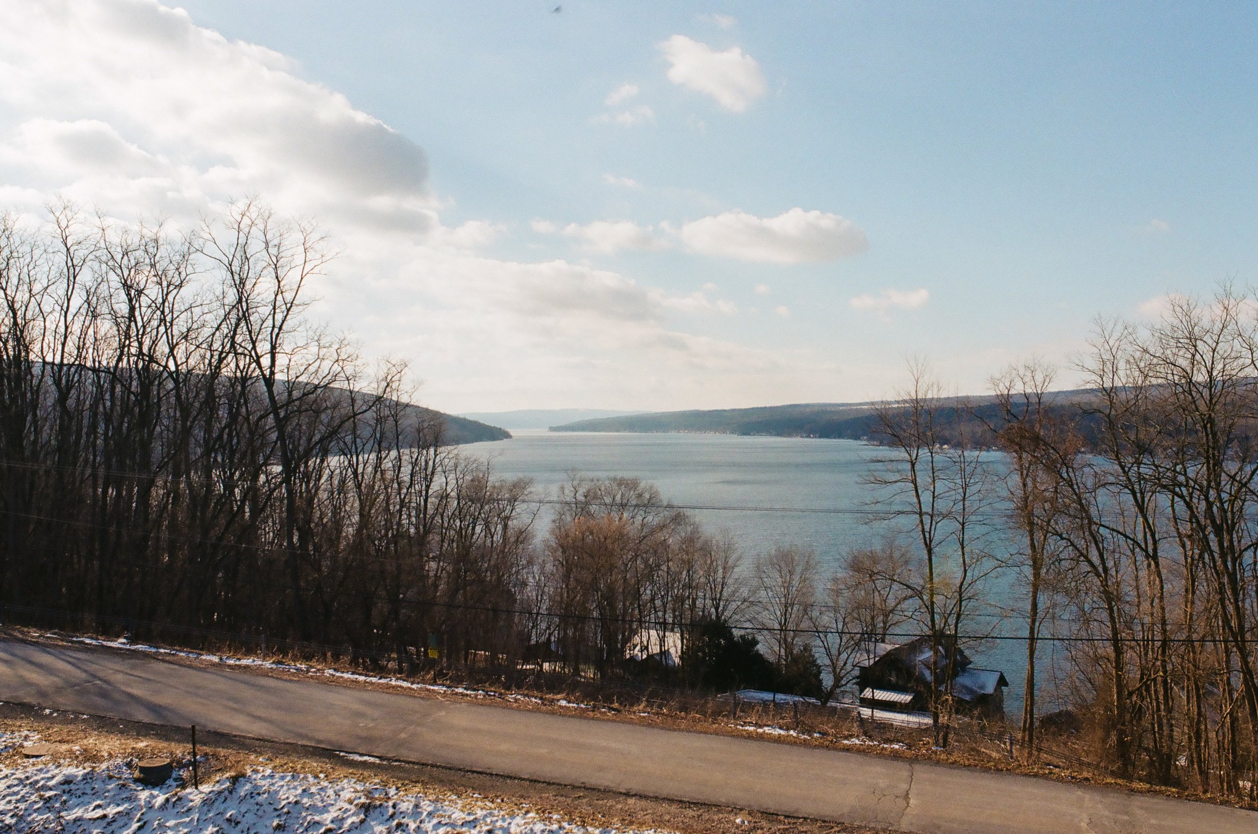 View of a wide lake with distant hills, leafless trees in the foreground, and a partly cloudy sky.