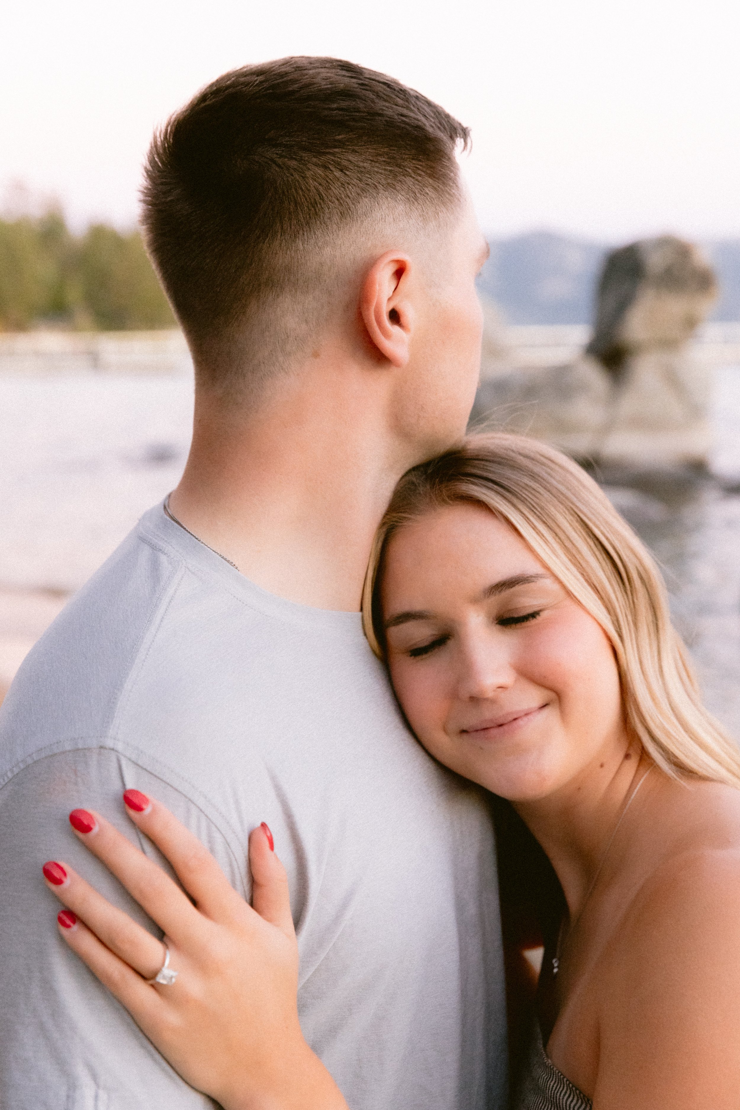 A young woman with long blonde hair embracing a man with short brown hair at the beach, both with their eyes closed and smiling softly.