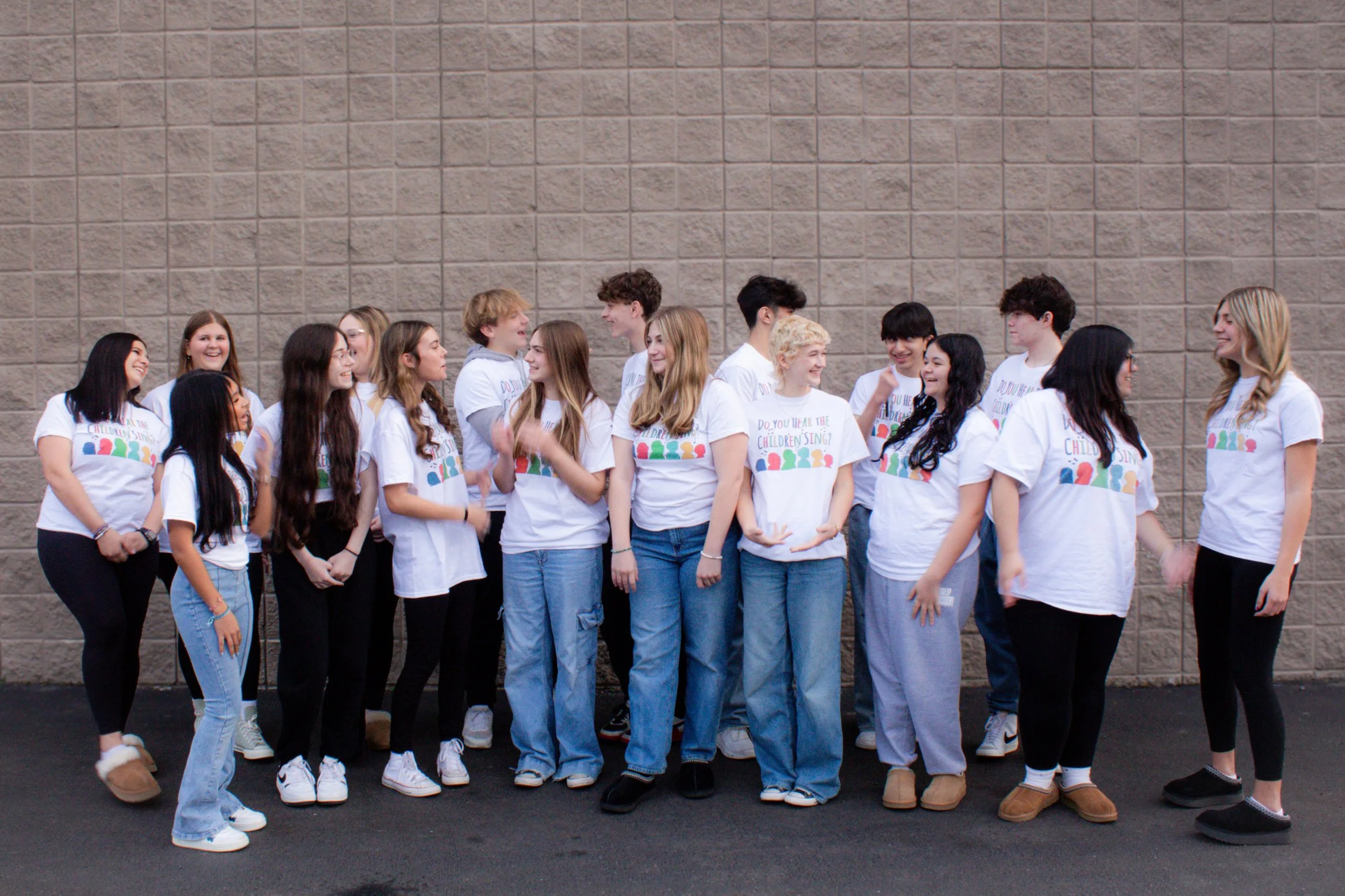 A group of teenagers standing and talking against a plain brick wall, wearing matching white T-shirts with colorful writing and graphics.