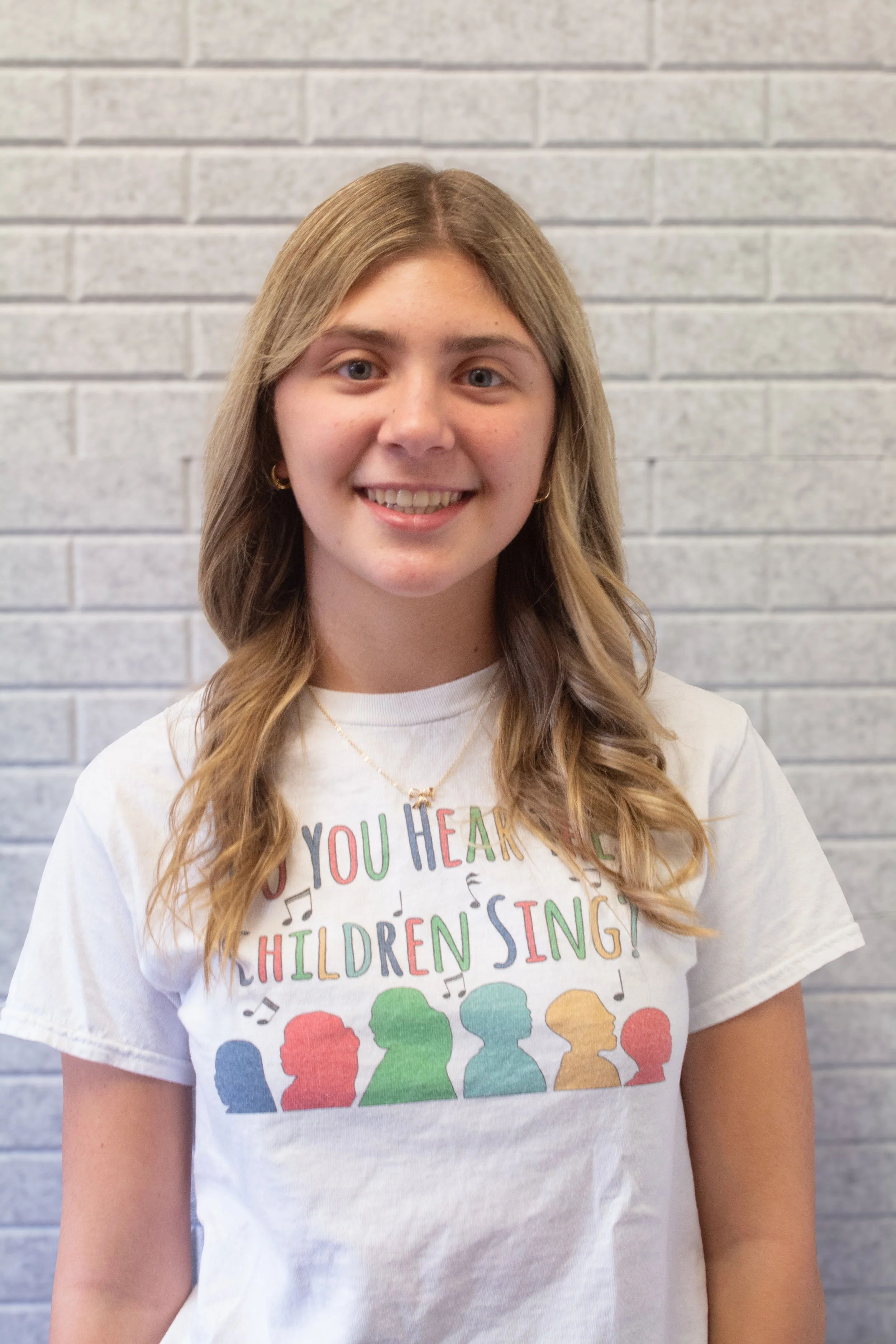 A young girl with blonde hair and blue eyes smiling, wearing a white T-shirt with colorful text and rainbow-colored silhouettes of children singing, standing in front of a light gray brick wall.