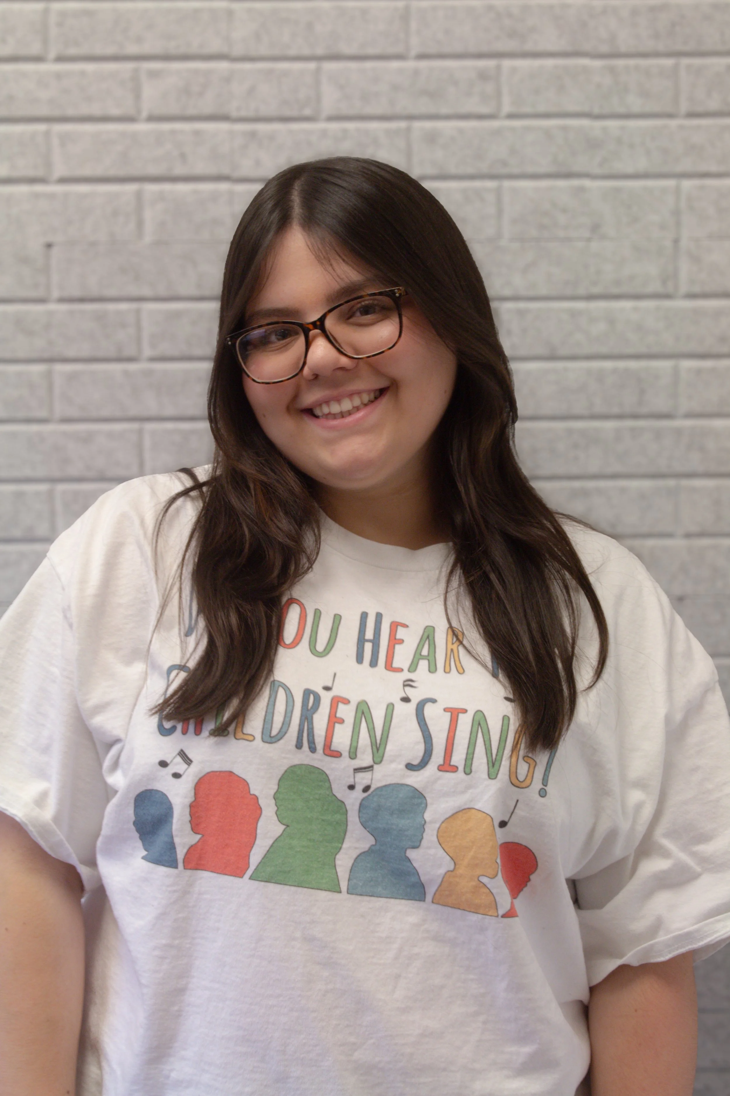 A young woman with glasses and long dark hair smiling, wearing a white T-shirt with colorful text and silhouettes of children, standing in front of a gray brick wall.