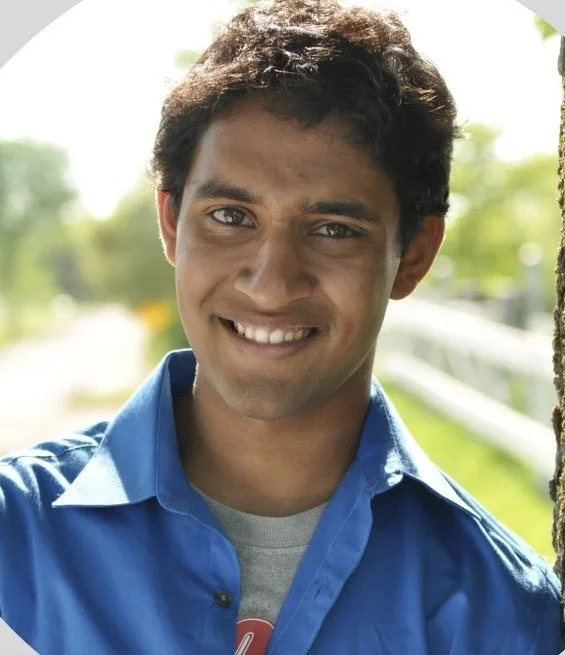 Young man smiling outdoors, wearing a blue shirt and gray t-shirt, with a blurred green background.