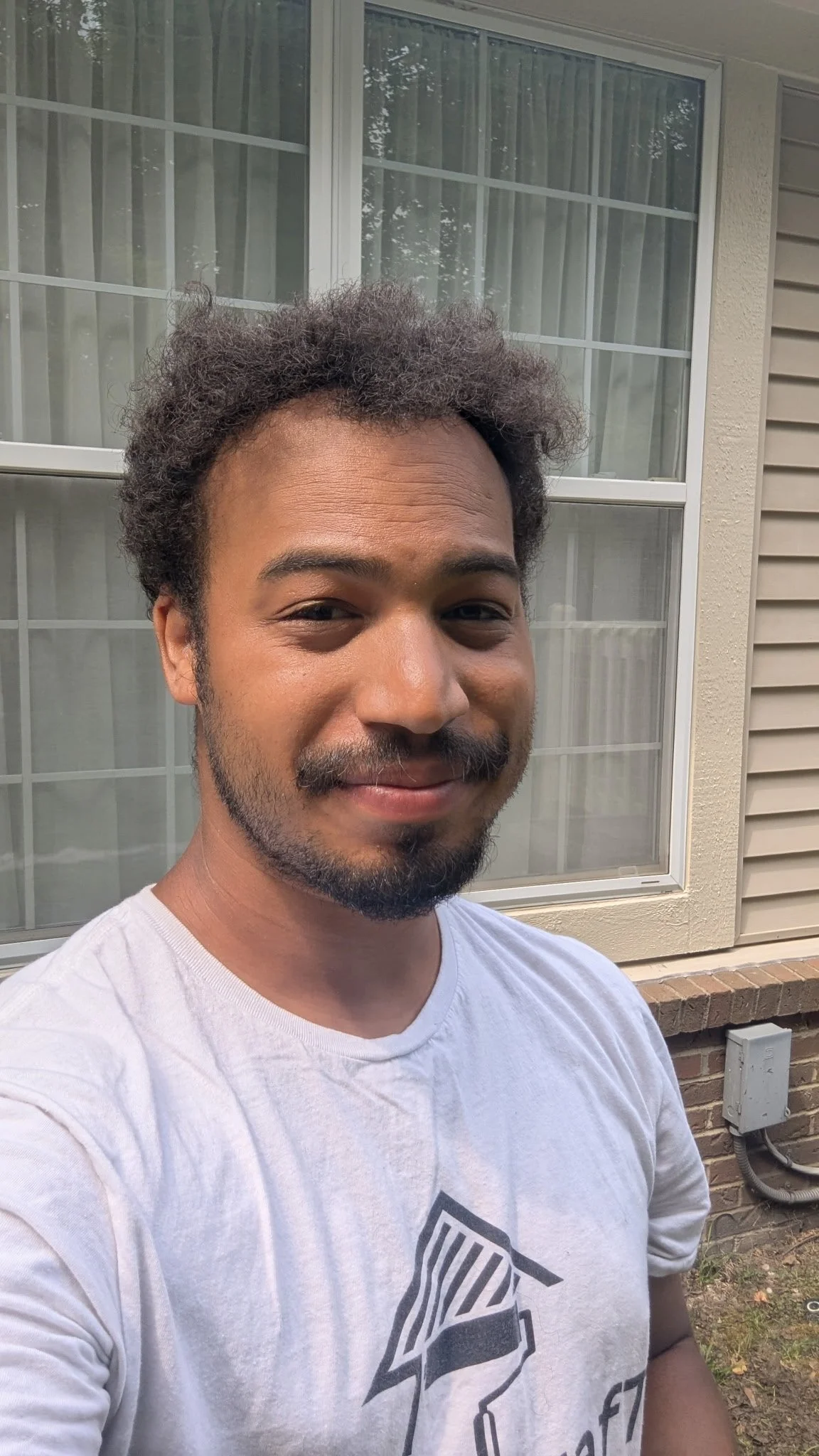 A young man with curly hair, a mustache, and beard, smiling outdoors in front of a window and beige house siding.