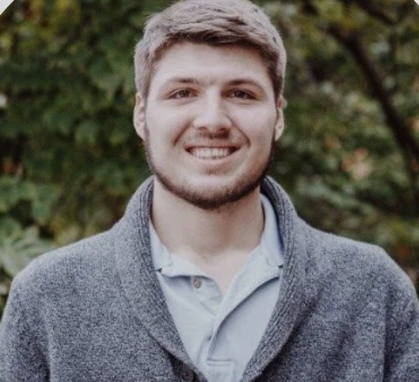 A young man with light brown hair and a beard, smiling, outdoors with green trees in the background, wearing a gray cardigan over a light-colored polo shirt.