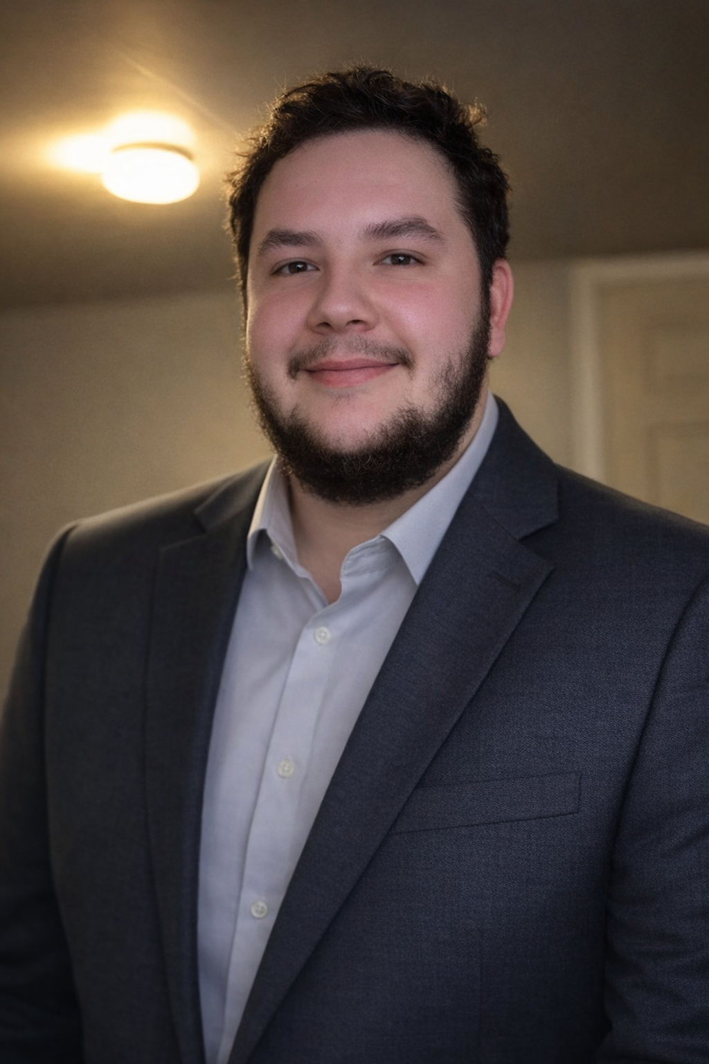 Young man with dark brown hair and beard, wearing a dark suit and white shirt, smiling in a professional headshot indoors with warm lighting.