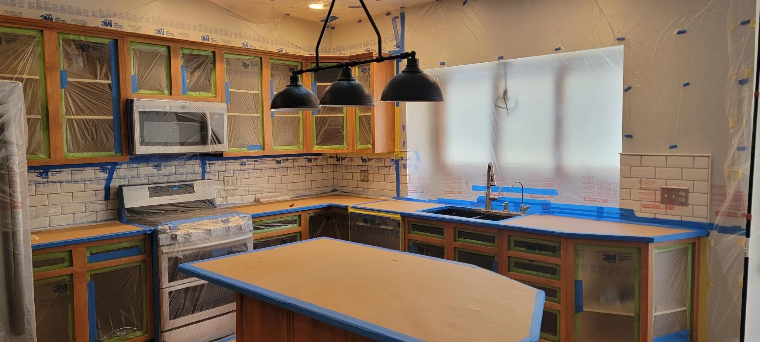 Kitchen under renovation with cabinets covered in plastic, a microwave, stove, and a sink with a window. Blue painter's tape is on cabinets and countertops, and unfinished walls with protective plastic covering.