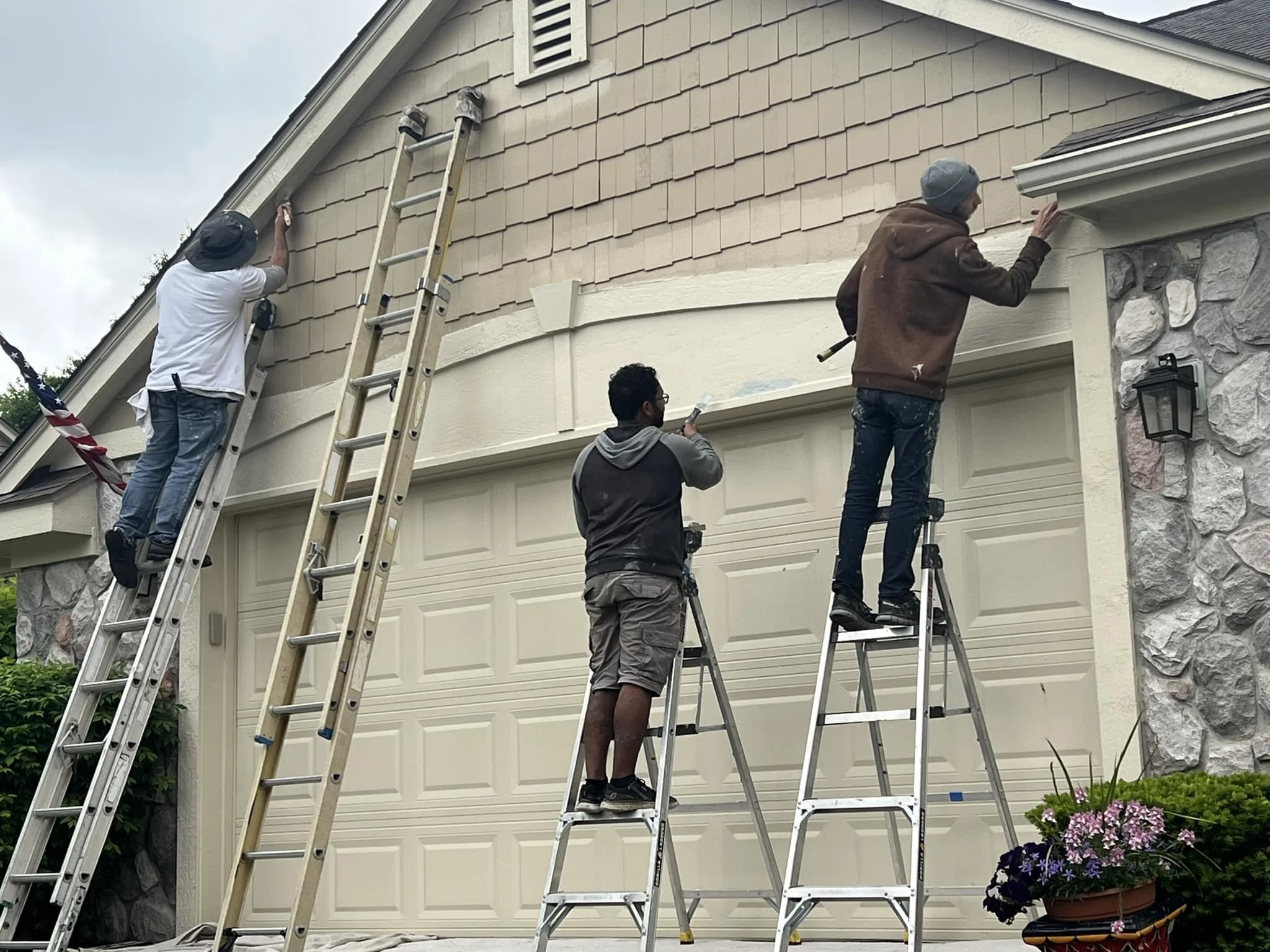 Four people working on painting the exterior of a house, with ladders and paint brushes, on a cloudy day.