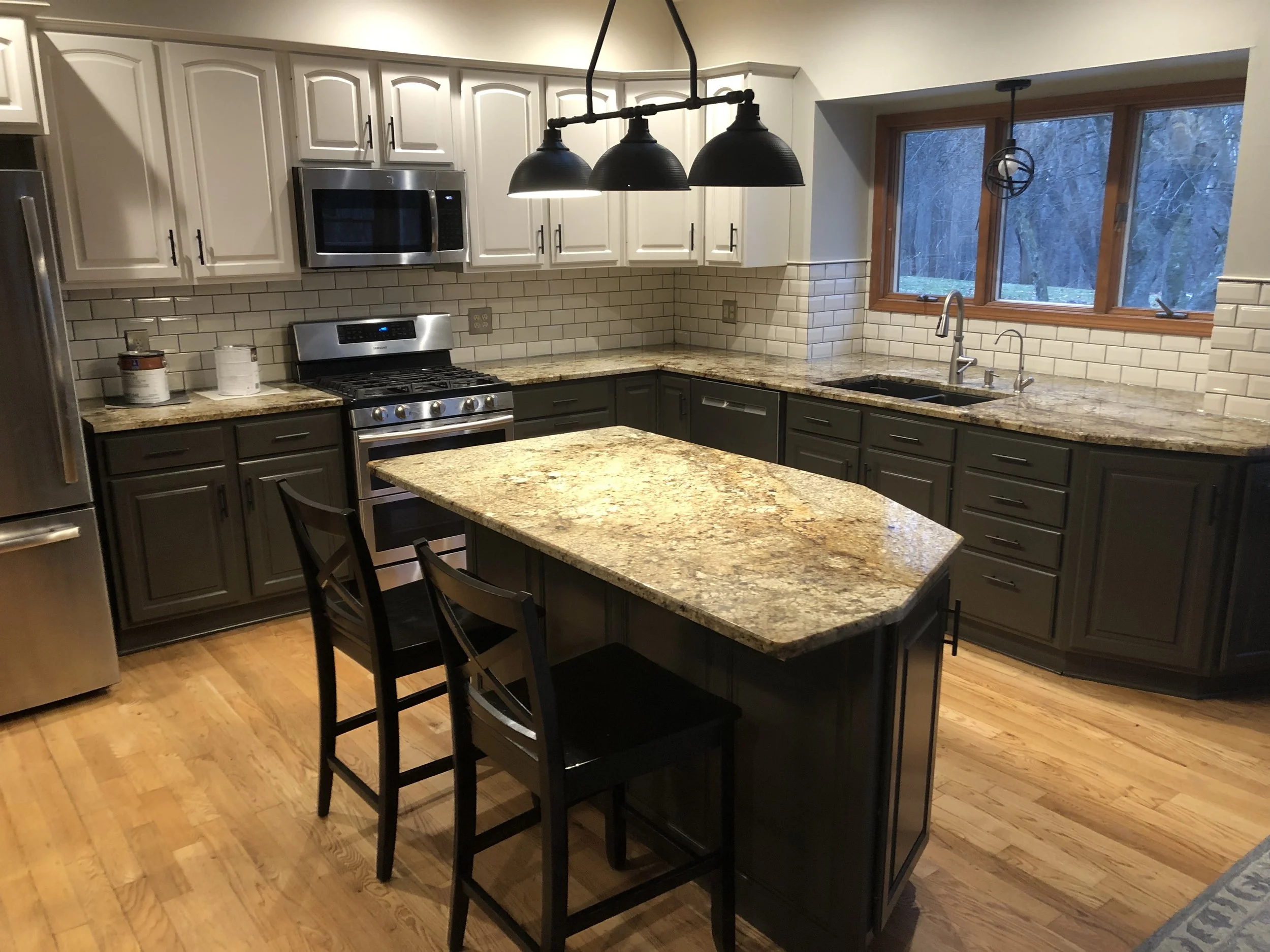 Kitchen with granite countertops, dark lower cabinets, light upper cabinets, a stainless steel refrigerator, oven, microwave, and a kitchen island with bar stools, a window above the sink, and a black pendant light fixture.