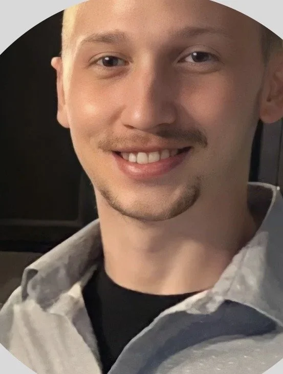 Close-up of a young man smiling, wearing a gray collared shirt, with short hair and facial hair, sitting in a vehicle.