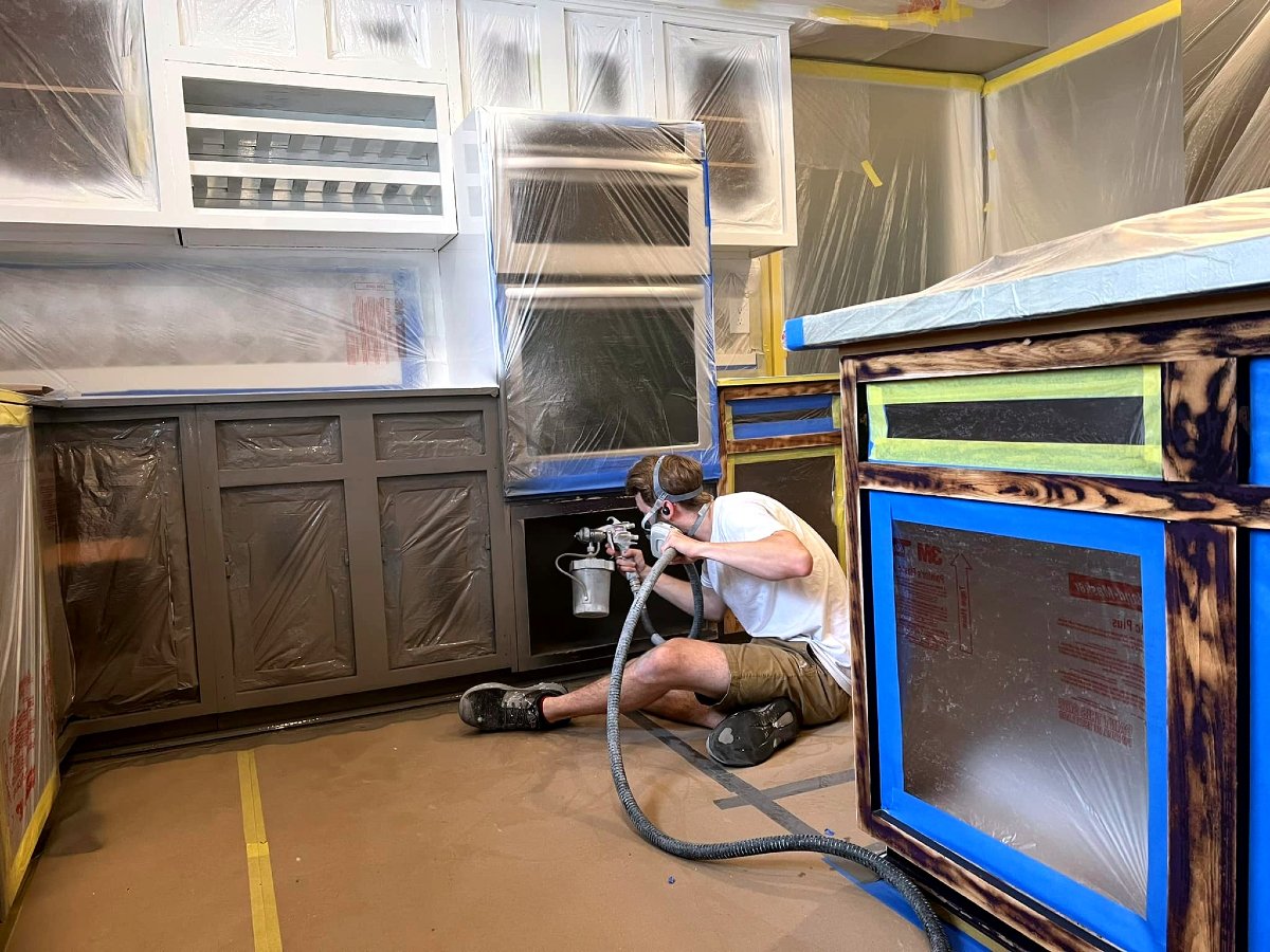 A person wearing safety goggles, a mask, and headphones is spray painting kitchen cabinets in a garage or workshop. The cabinets are covered with plastic and painter's tape for protection.