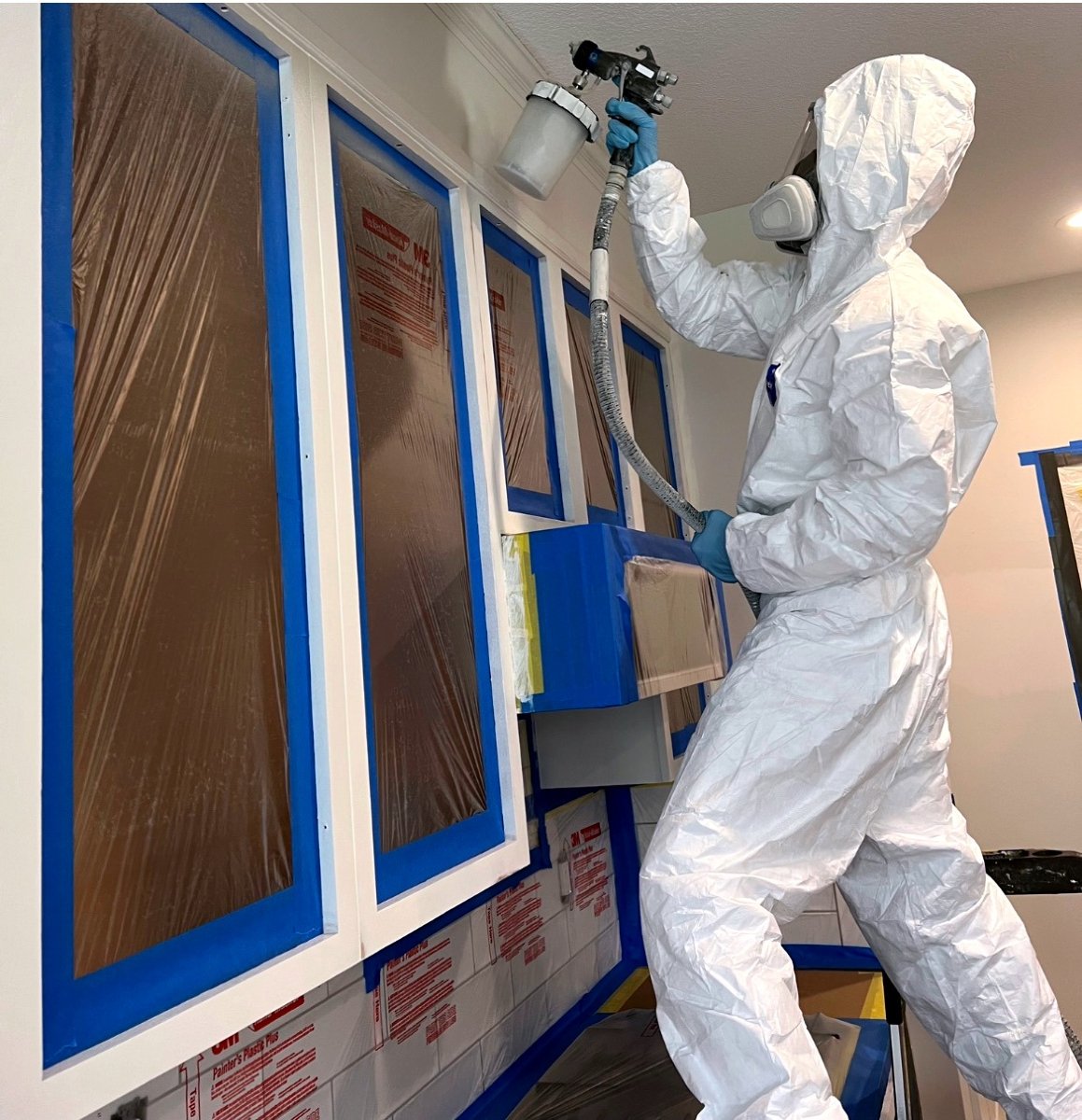 A worker in protective gear spray painting kitchen cabinets.