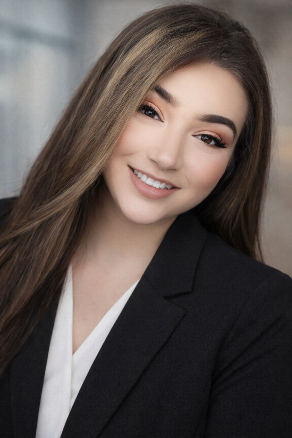 A young woman with long brown hair and makeup, wearing a black blazer and white blouse, smiling at the camera.