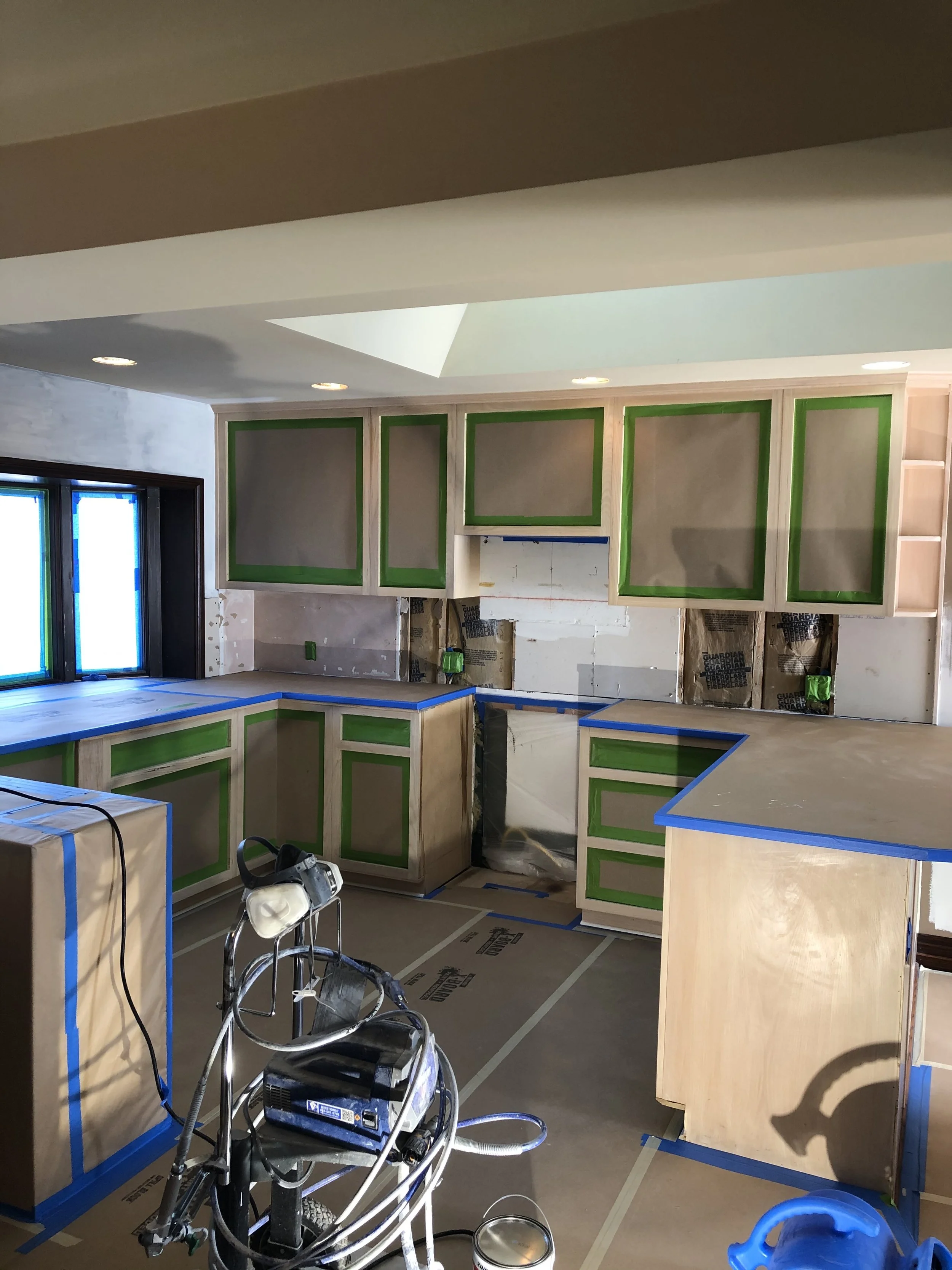 Kitchen under renovation with cabinets being painted, blue painter's tape, construction tools, and covered surfaces.