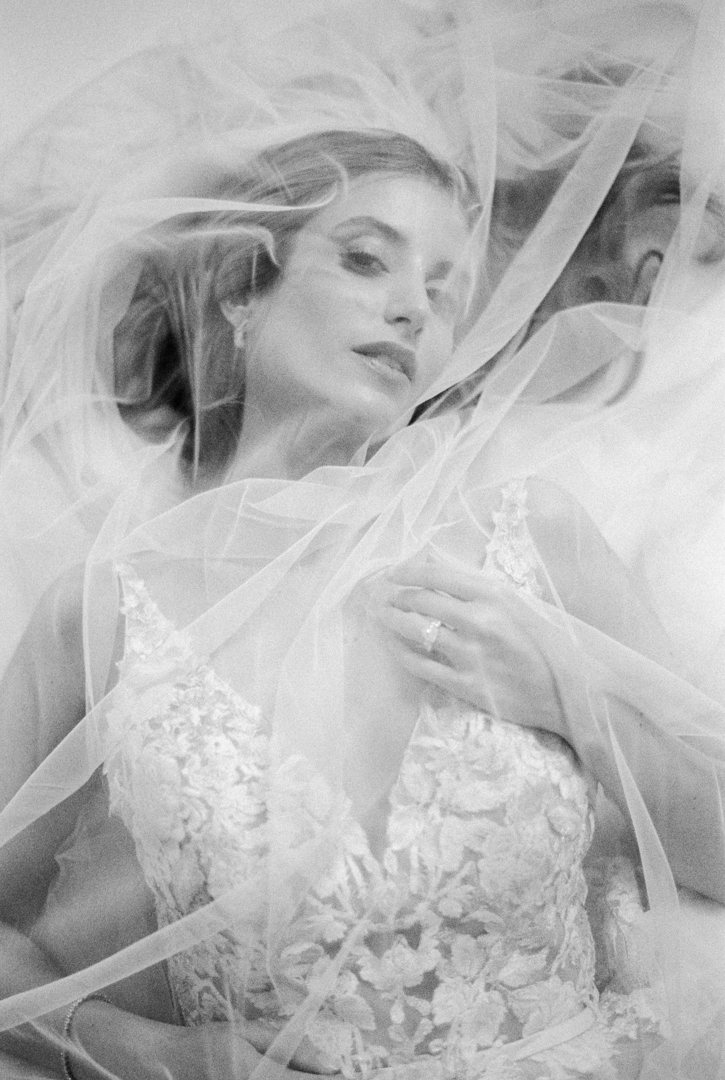 Black and white photo of a woman in a wedding dress lying on her back, wrapped in a sheer fabric.